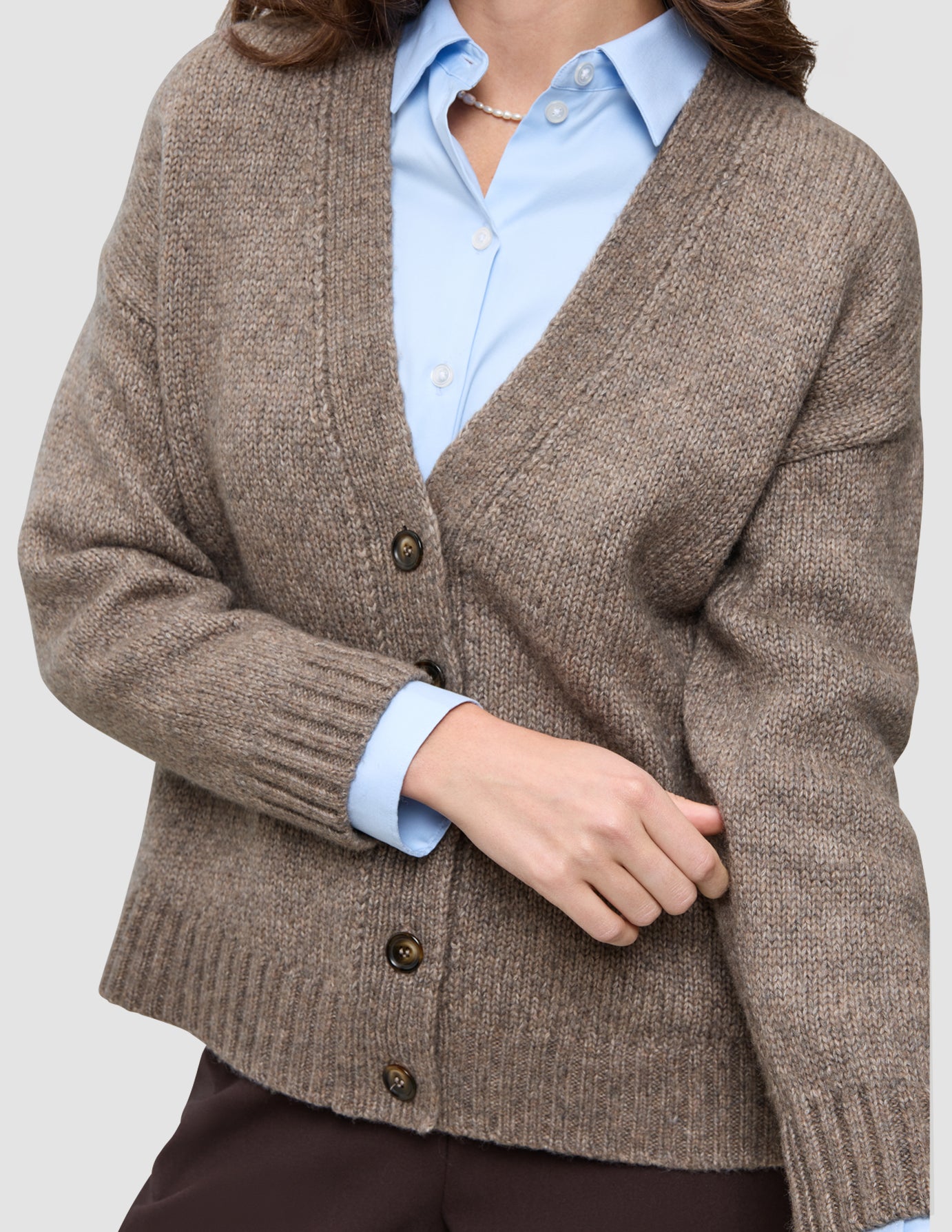 En tykk, myk ullstrikket damekardigan i taupe-melange, båret over en lyseblå skjorte med knapper.