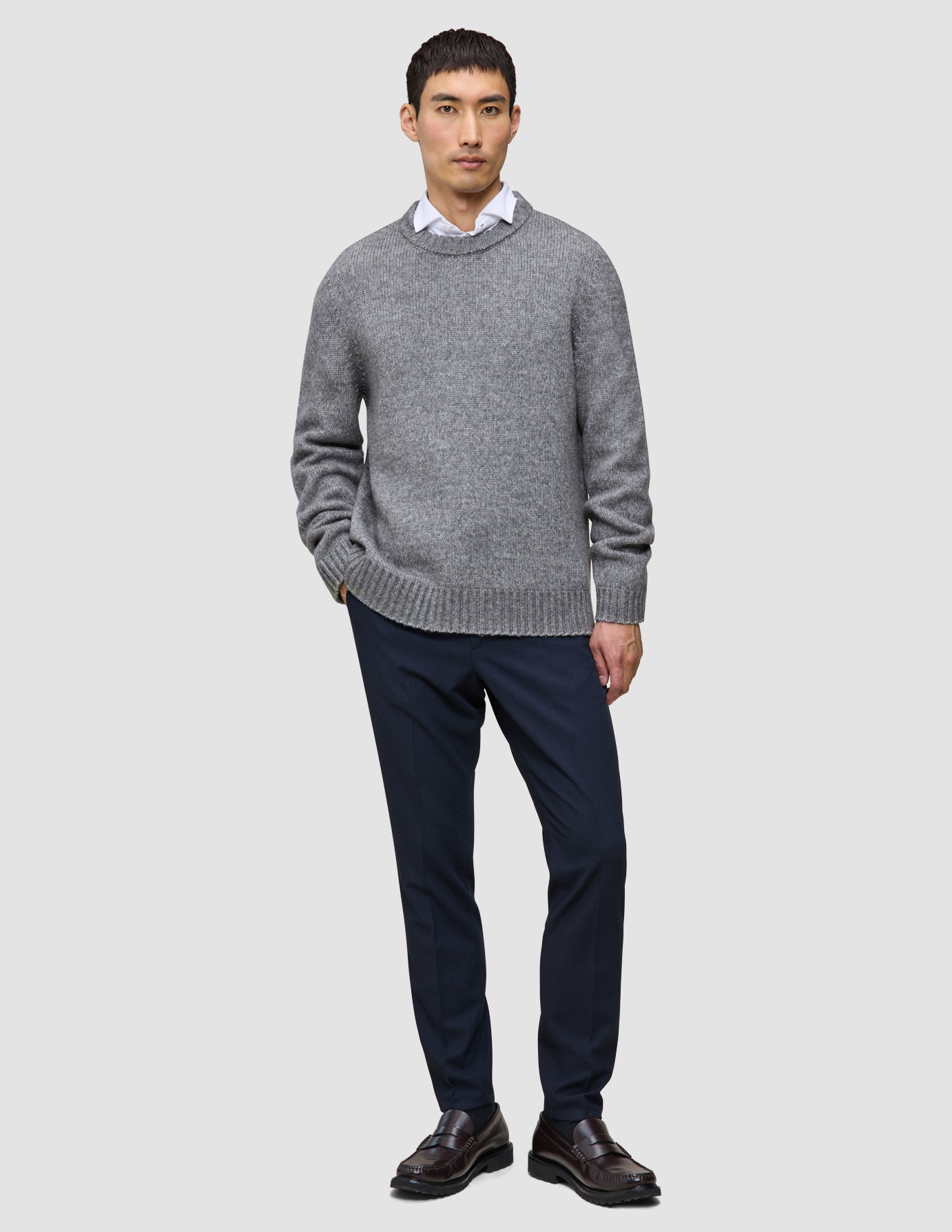 En mann i en tykk, myk ullgenser med rund hals i fargen Glacier Grey, over en hvit skjorte med krage, marineblå bukser og mørke loafers.