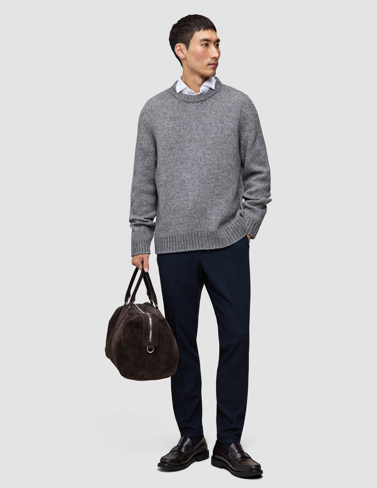 En mann i en tykk, myk ullgenser med rund hals i fargen Glacier Grey, hvit skjorte med krage, mørke bukser og loafers, som holder en reiseveske.