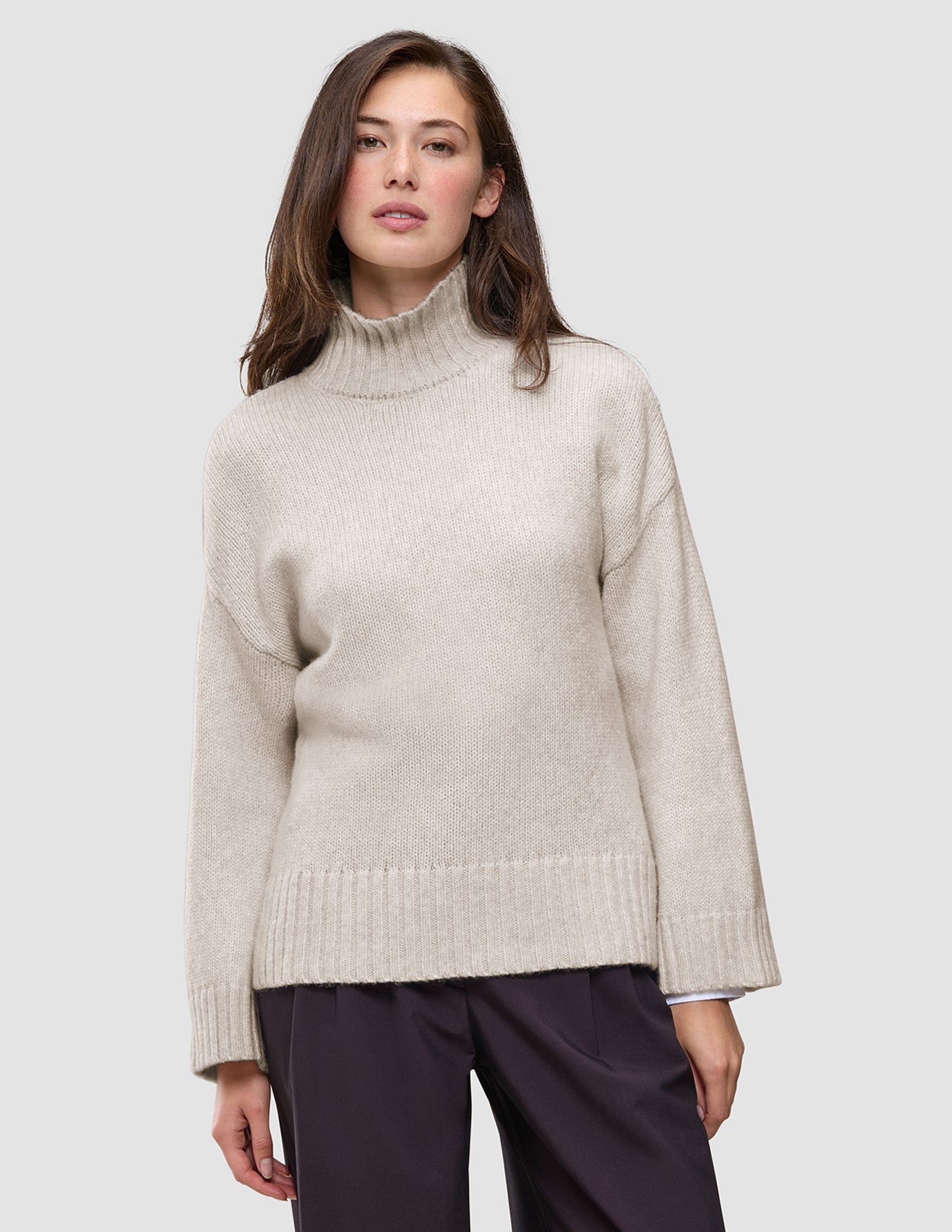 En kvinne har på seg en beige damegenser i tykk, myk ull med høyhals og Sugar Melange-mønster, med nedsenkede skuldre.