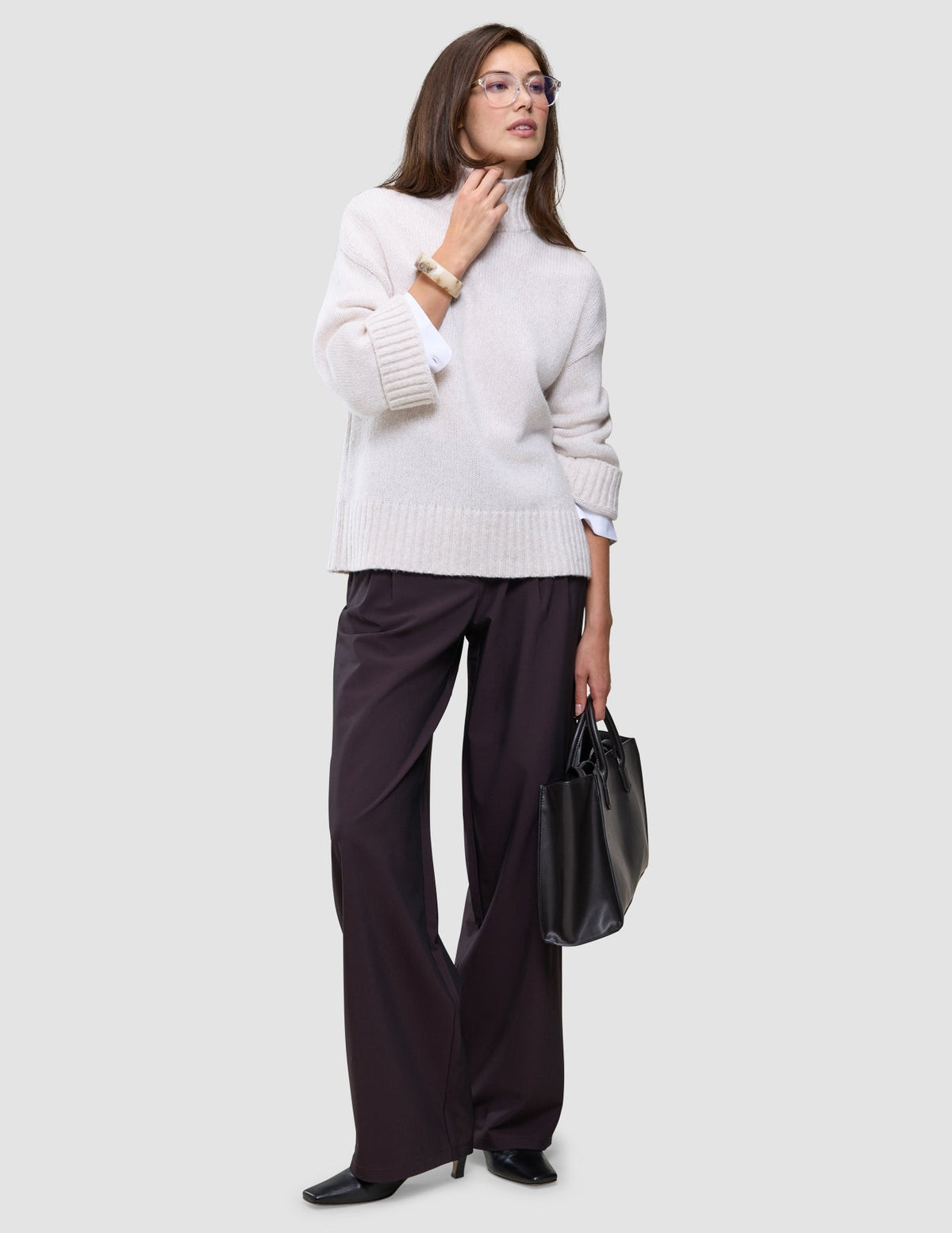 En kvinne kledd i en Women’s Chunky Soft Wool Knit Turtleneck Sugar Melange og mørke bukser med vide ben, som holder en svart håndveske.