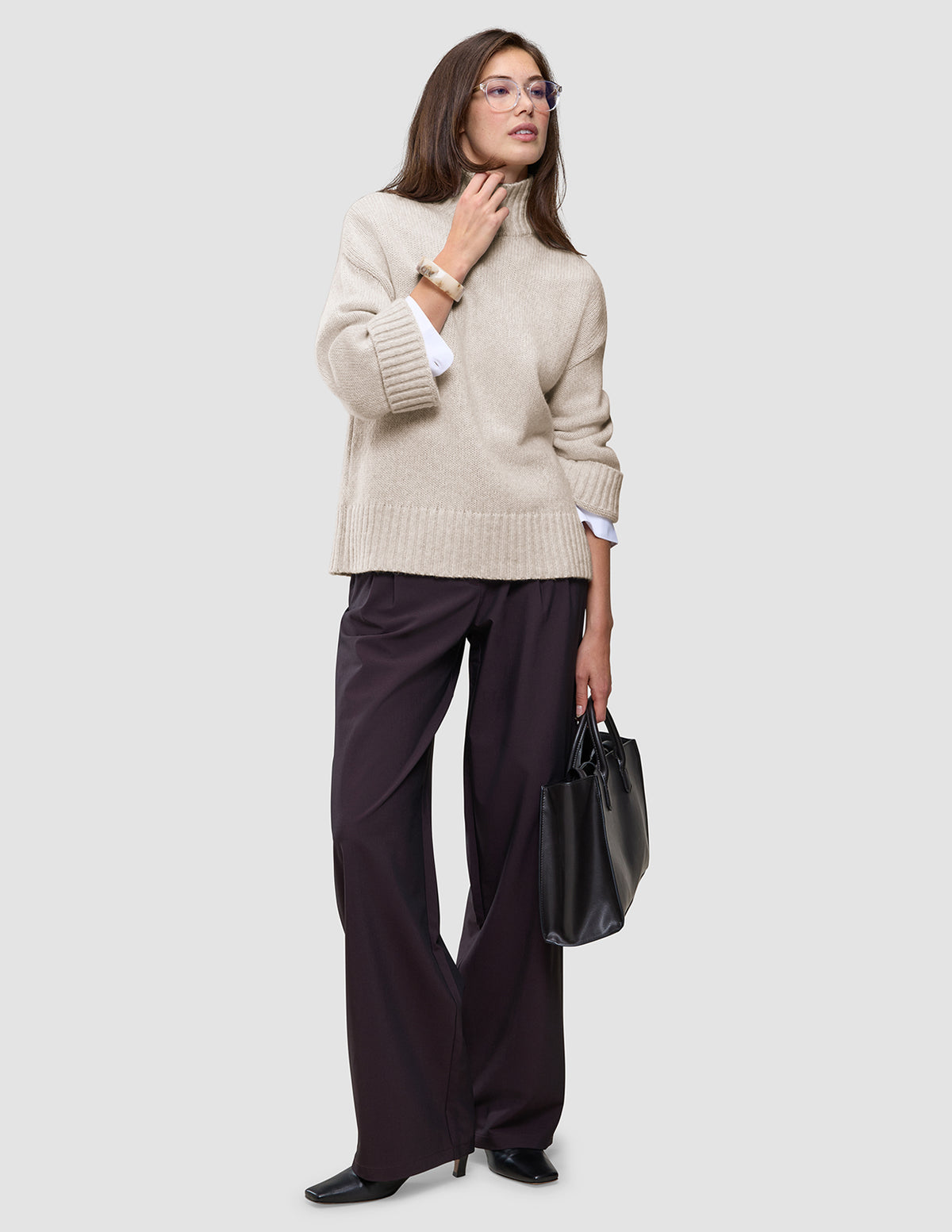 En kvinne har på seg en Women’s Chunky Soft Wool Knit Turtleneck Sugar Melange over en hvit skjorte og mørke bukser med vide ben.