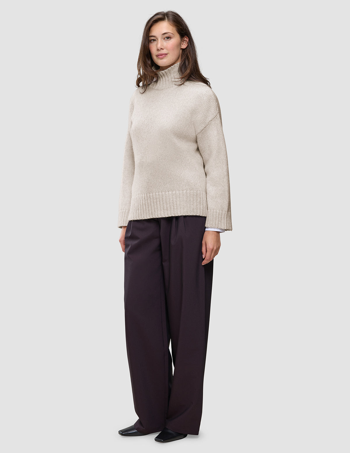 En kvinne kledd i en beige Women’s Chunky Soft Wool Knit Turtleneck Sugar Melange-genser og mørke bukser med vide ben.