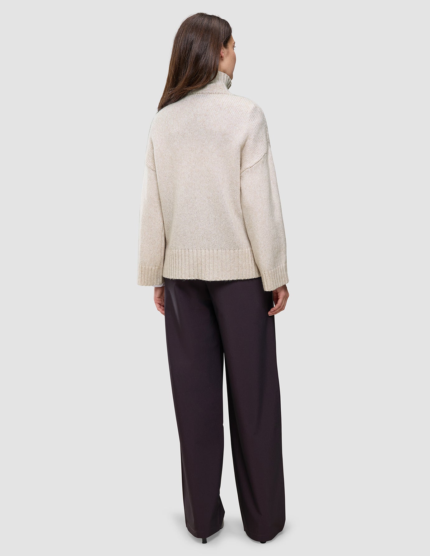 En kvinne sett bakfra, kledd i en tykk, myk ullstrikket turtleneck for kvinner i fargen Sugar Melange og mørke bukser med vide ben.