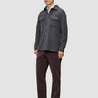 En mann har på seg en mørkegrå, vanlig dobbeltsidig ullskjorte over en hvit skjorte, kombinert med mørkebrune bukser og loafers.