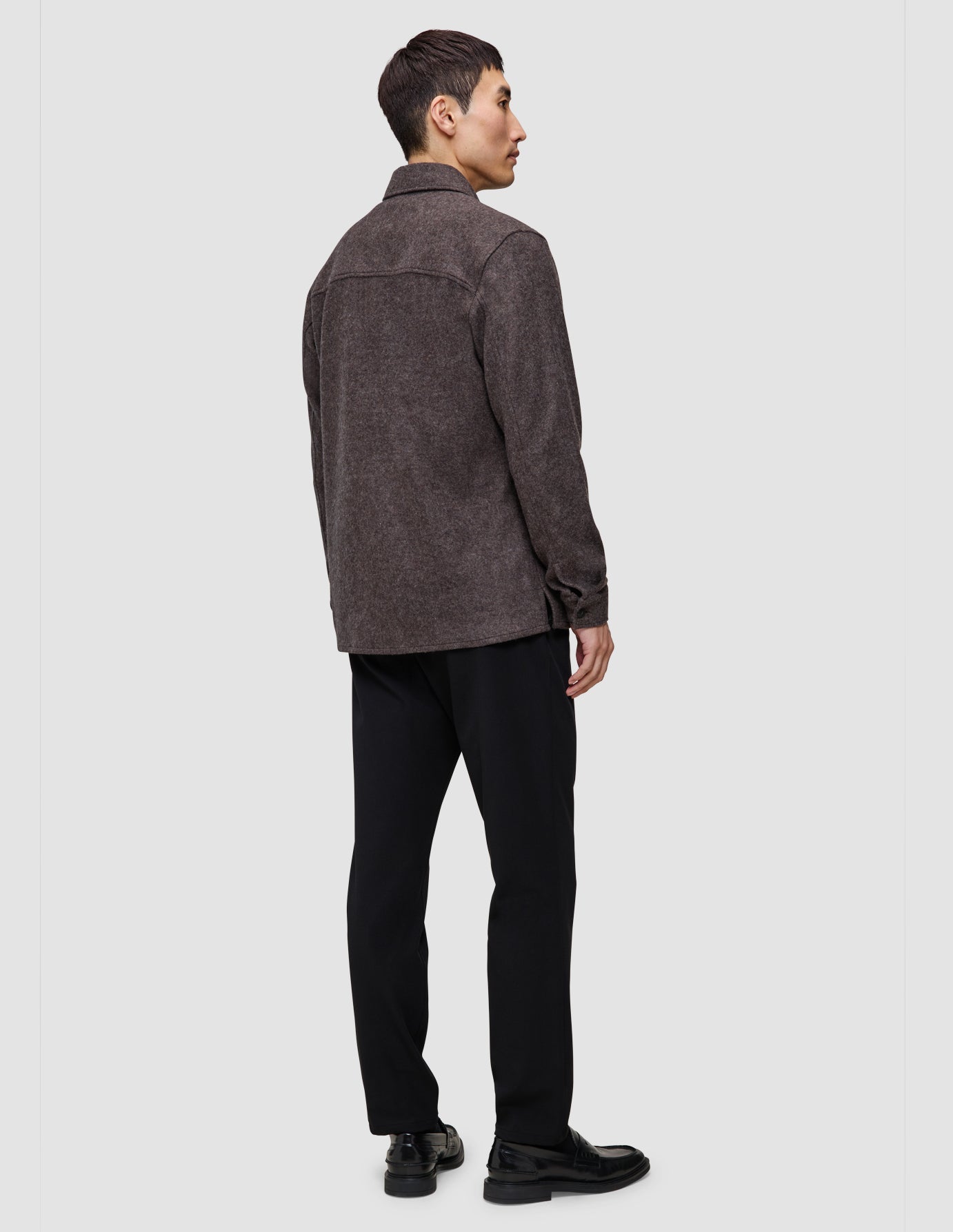 En person sett bakfra, kledd i en dobbeltlags ullskjorte i sadelbrunt, svarte bukser og svarte loafers.