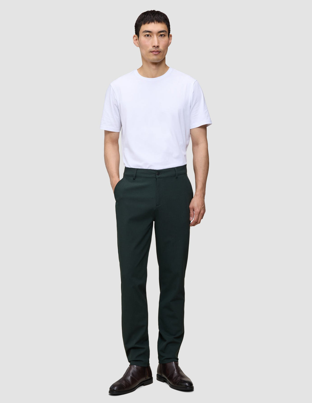 En mann har på seg en hvit T-skjorte, Ease Pants Regular Northern Pine og mørke skinnsko i en minimalistisk skandinavisk stil.