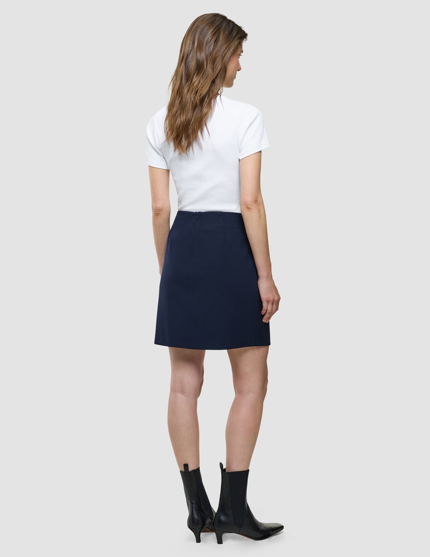 En kvinne sett bakfra, kledd i en hvit T-skjorte, et «Women Ease Short Skirt Blue Nights»-skjørt og svarte ankelstøvler.