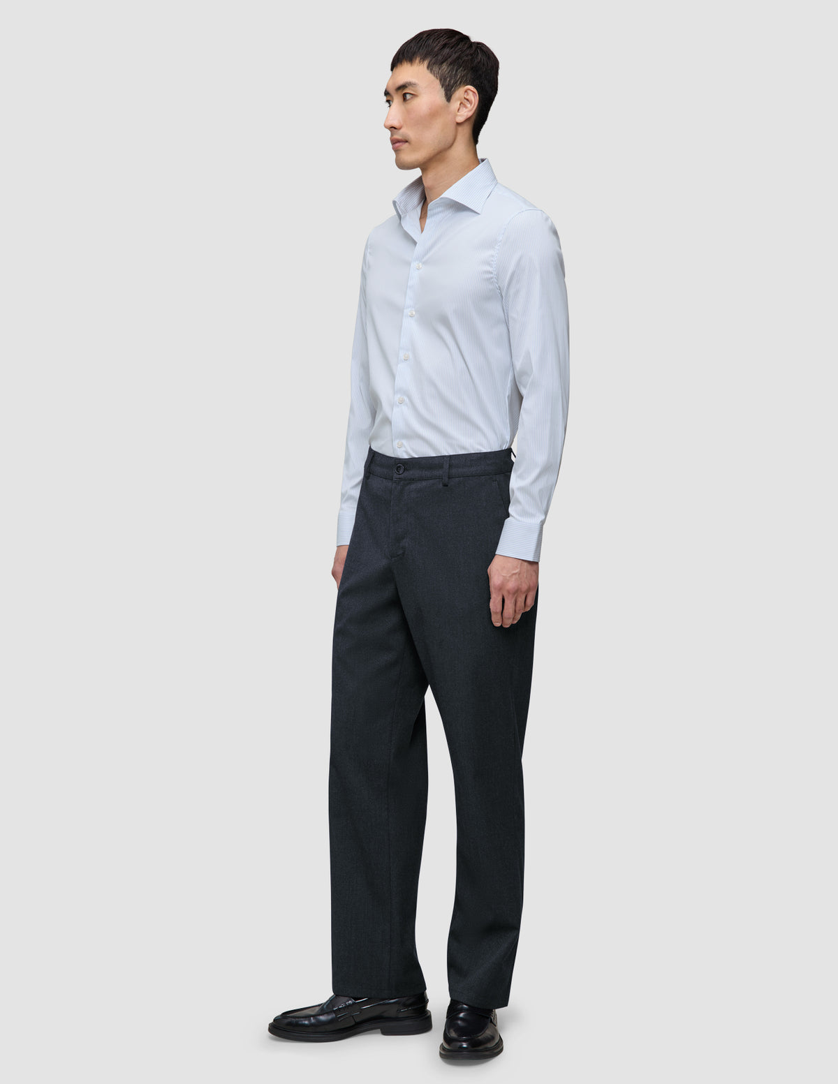 En mann i en Elevated Shirt Regular Classic Blue Striped – en lysblå skjorte med knapper – som er stappet ned i grå bukser og dressko.
