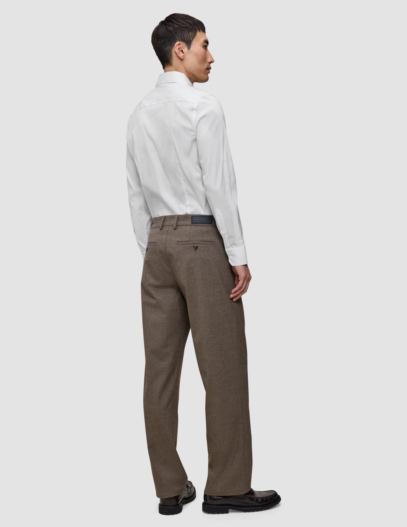 En mann sett bakfra har på seg en hvit, smal Elevated Shirt som er stappet ned i brune bukser og mørke skinnloafers.