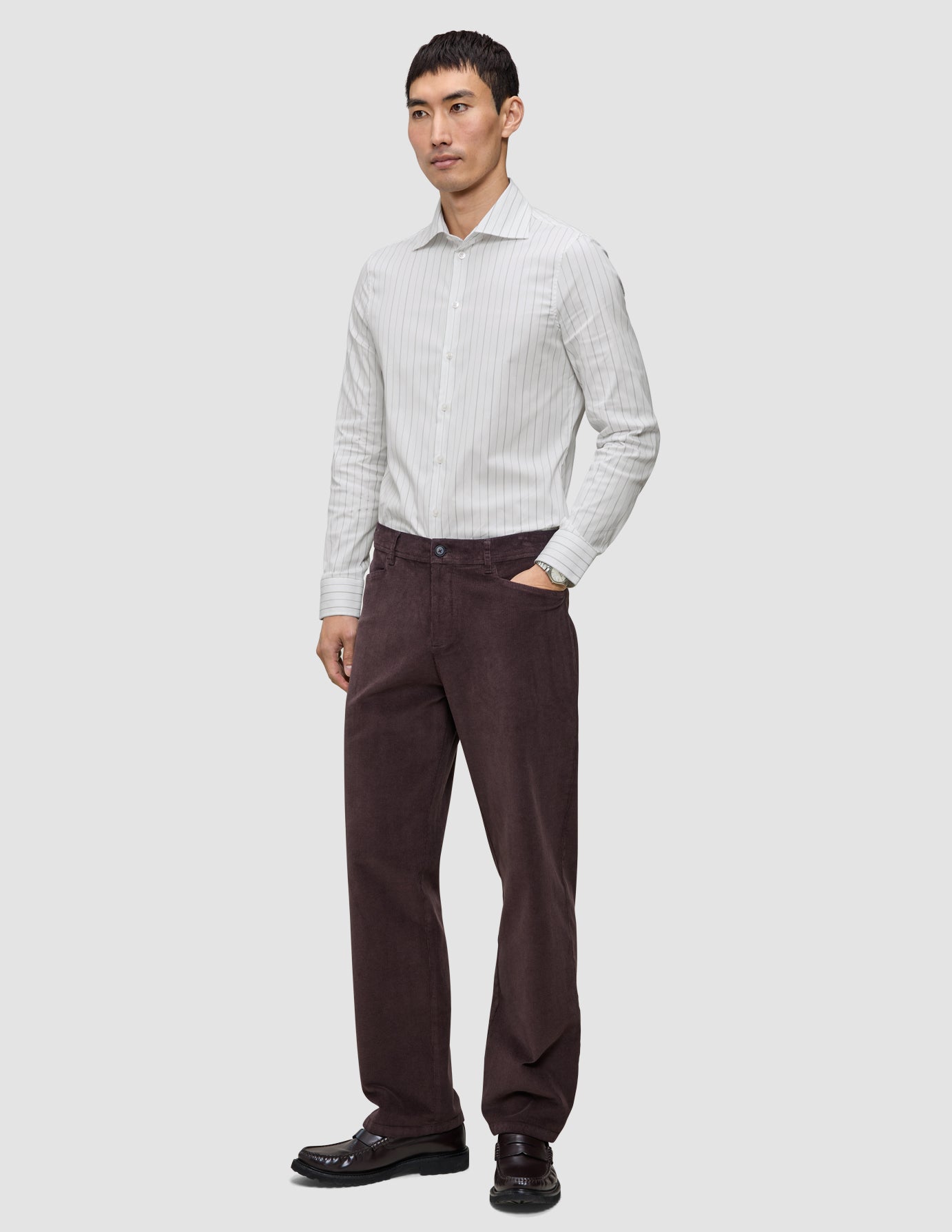 En mann i en Elevated Shirt Slim med lysegrå striper, mørkebrune bukser og mørke loafers.