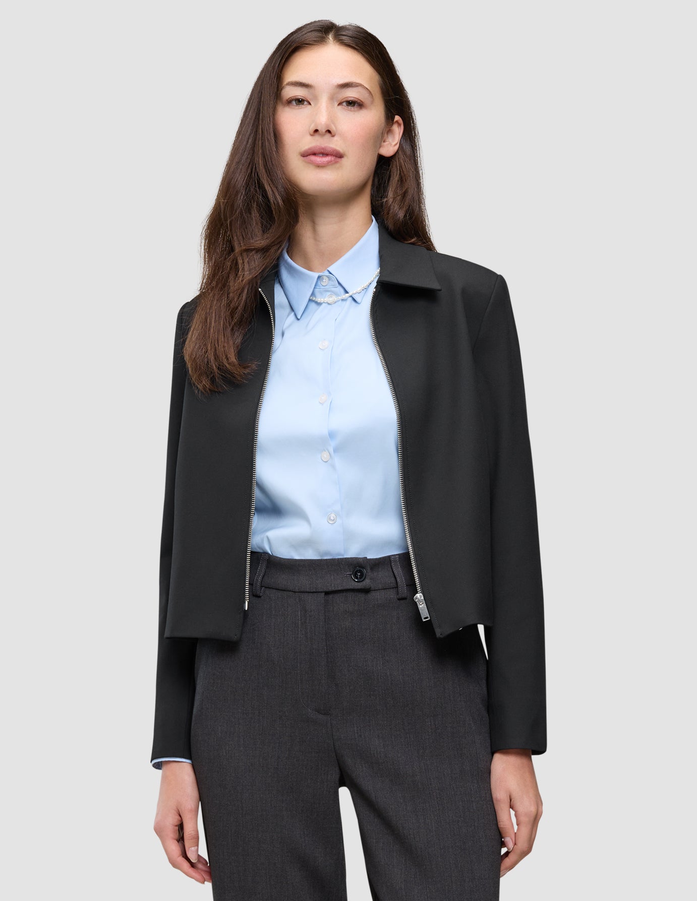 En kvinne har på seg en svart Essential Short Zip Jacket over en lyseblå skjorte med knapper og grå bukser.