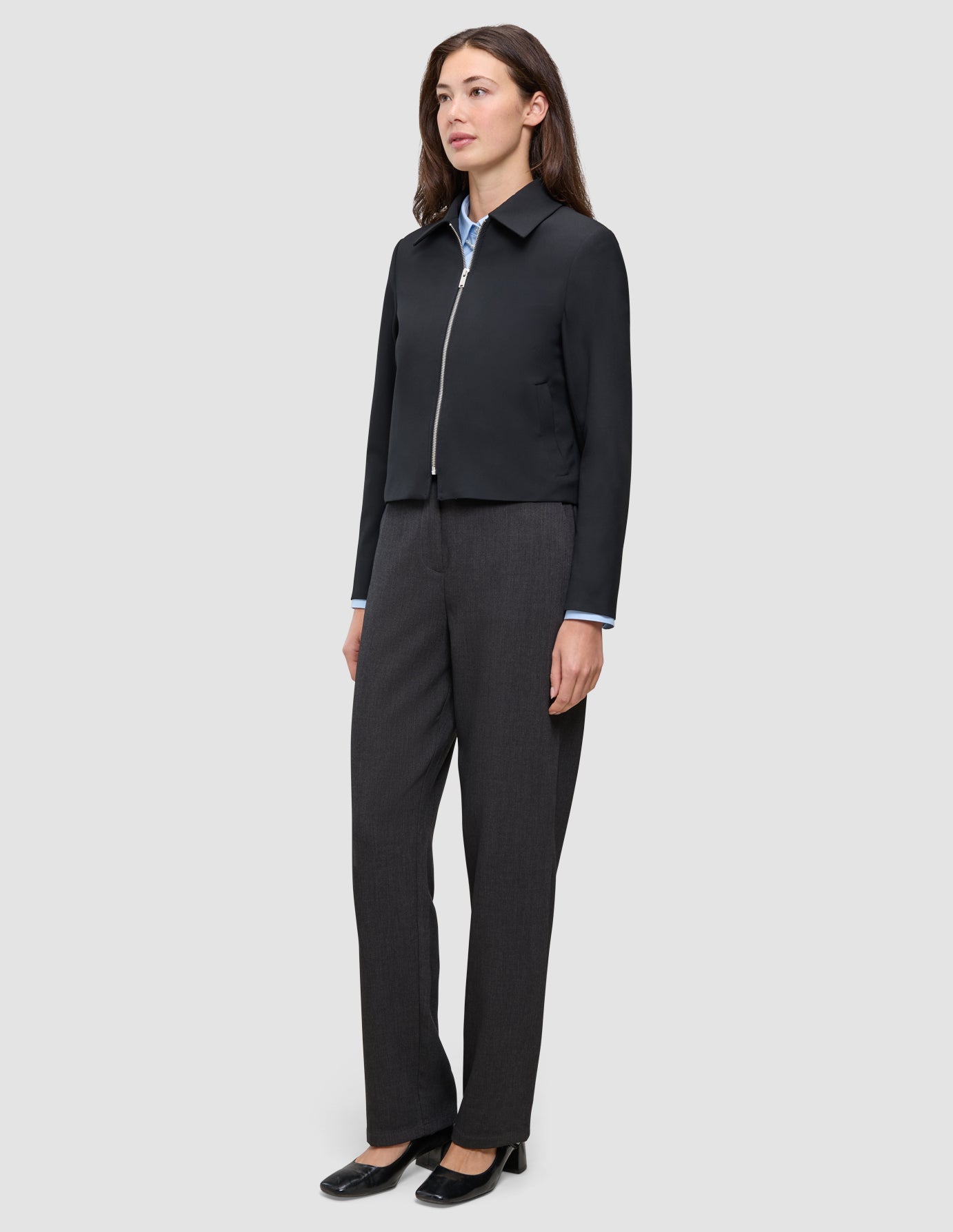 En kvinne har på seg en svart Essential Short Zip Jacket over en lyseblå skjorte med krage og mørkegrå bukser.