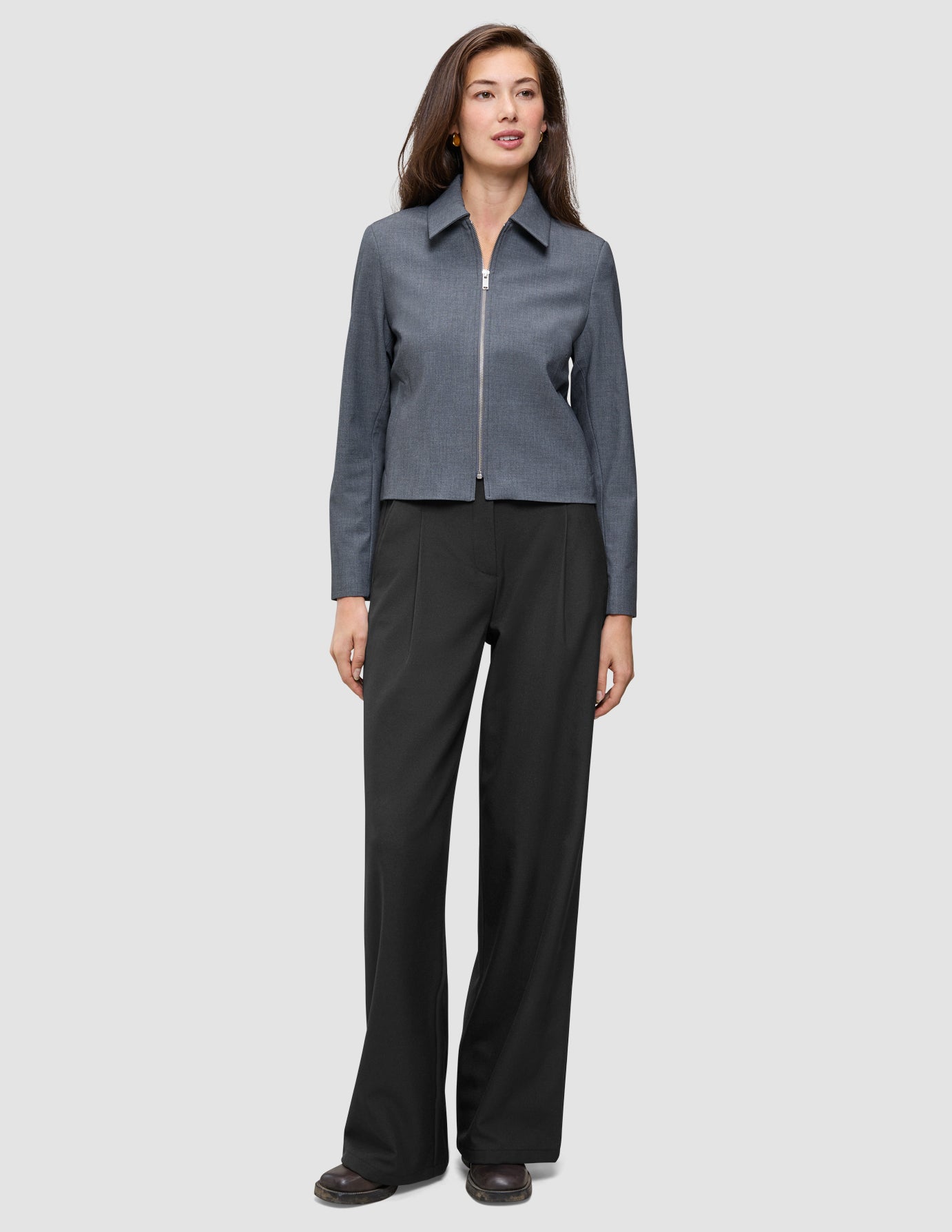 En kvinne har på seg en Essential Short Zip Jacket Dark Grey Melange – en minimalistisk jakke med krage og glidelås foran – samt svarte bukser.