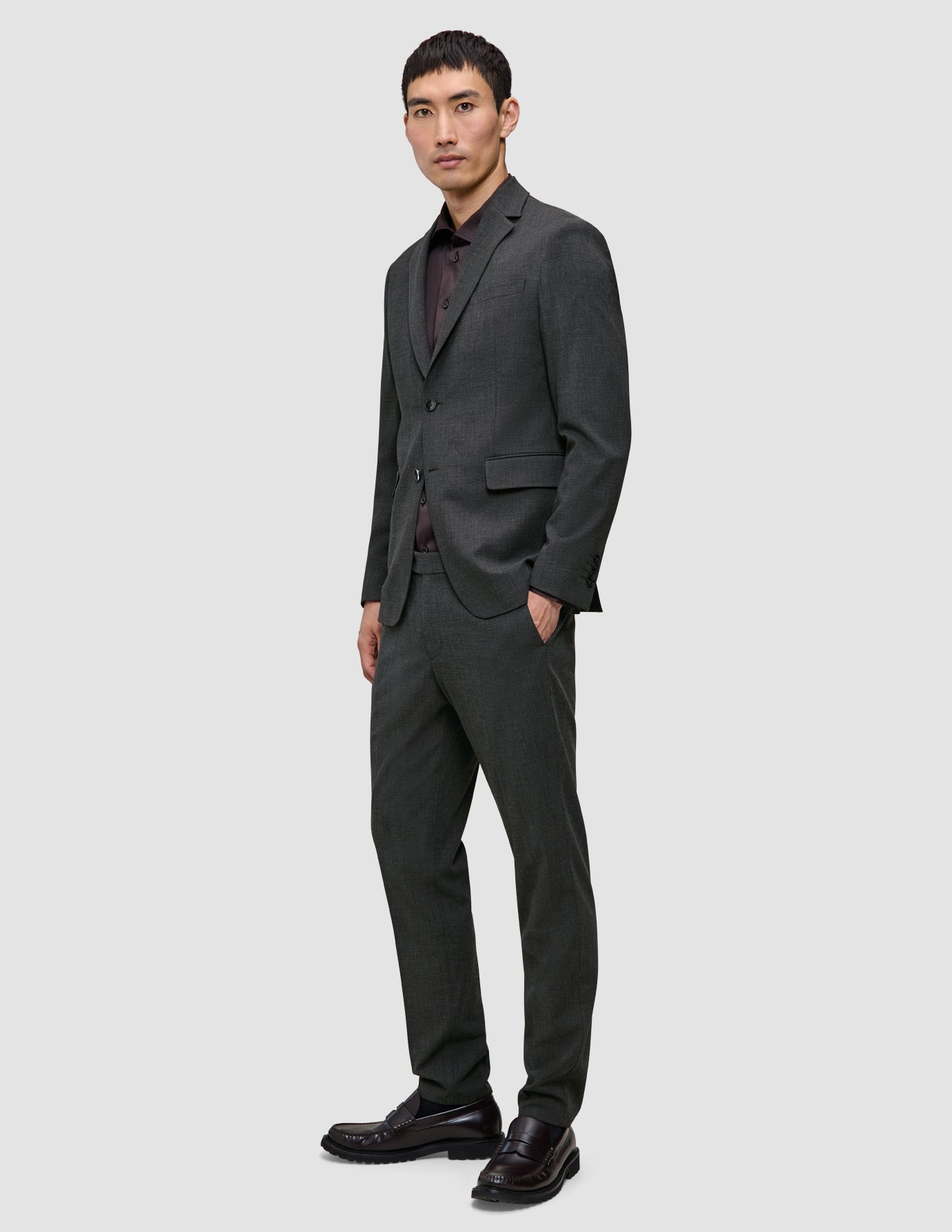 En mann kledd i en Motion Blazer Regular i fargen Pepper Grey, en rødbrun skjorte og mørke loafers står med den ene hånden i lommen.