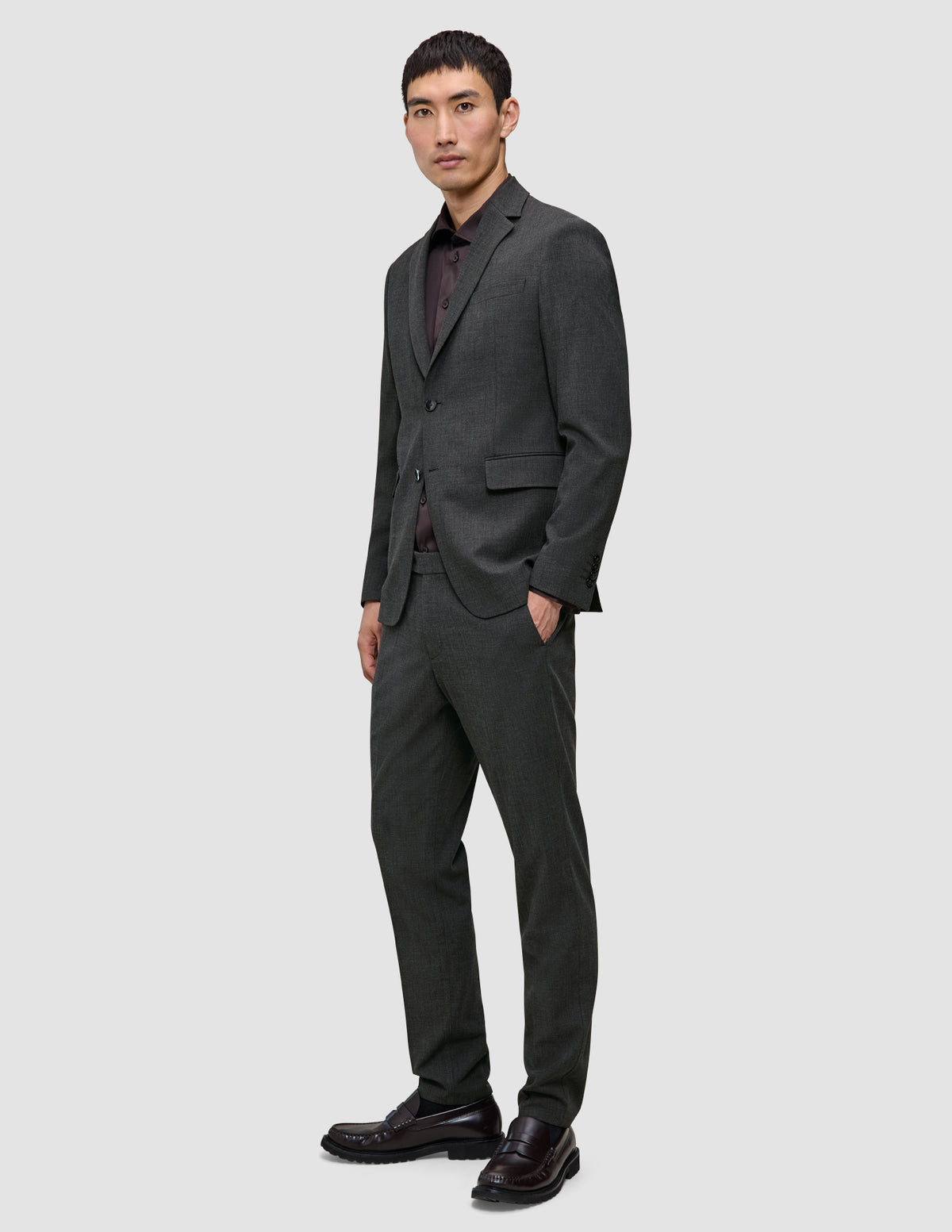 En mann kledd i en Motion Blazer Regular i fargen Pepper Grey, en rødbrun skjorte og mørke loafers står med den ene hånden i lommen.