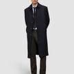 En mann i svart overfrakk, dressvest, skjorte og slips har på seg Motion Suit Pants Regular Cognac med hendene bak ryggen.