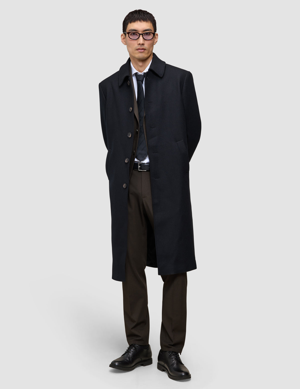En mann i svart overfrakk, dressvest, skjorte og slips har på seg Motion Suit Pants Regular Cognac med hendene bak ryggen.