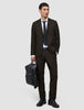 En mann kledd i Motion Suit-bukser i fargen Regular Cognac, en hvit skjorte og et slips, som holder en svart ryggsekk.