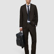 En mann kledd i Motion Suit-bukser i fargen Regular Cognac, en hvit skjorte og et slips, som holder en svart ryggsekk.