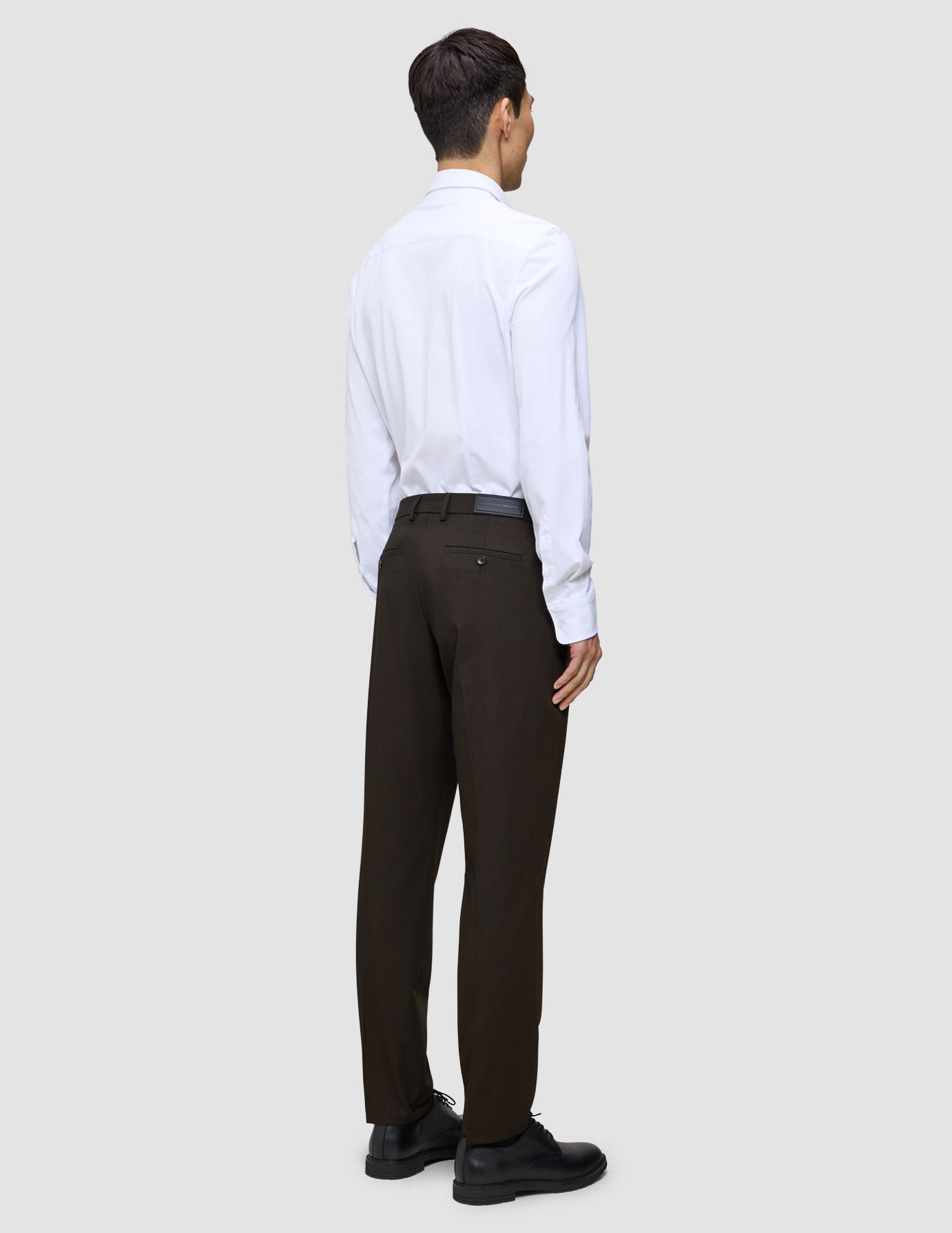 En person sett bakfra, kledd i en hvit skjorte, Motion Suit-bukser i Regular-passform i fargen Cognac og svarte dressko.