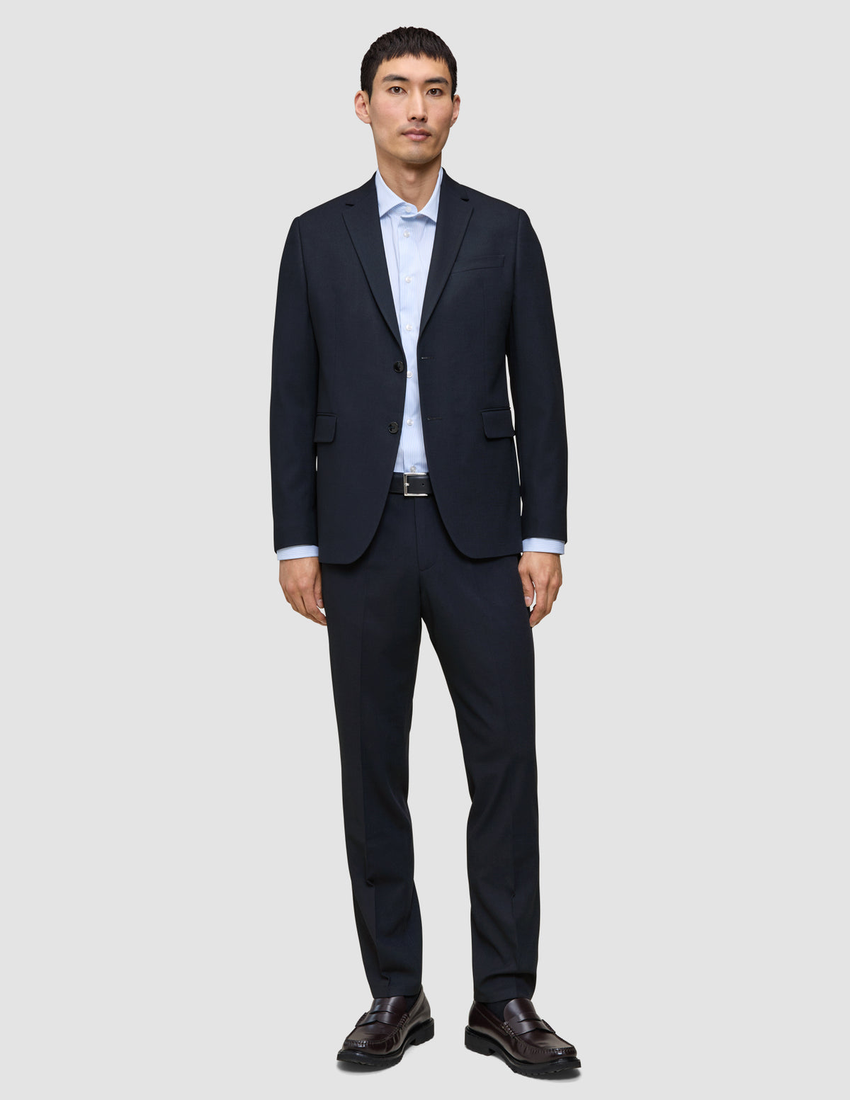En mann kledd i Motion Suit-bukser i størrelse Regular i fargen Midnight Blue, med en lyseblå skjorte og mørke skinnloafers.