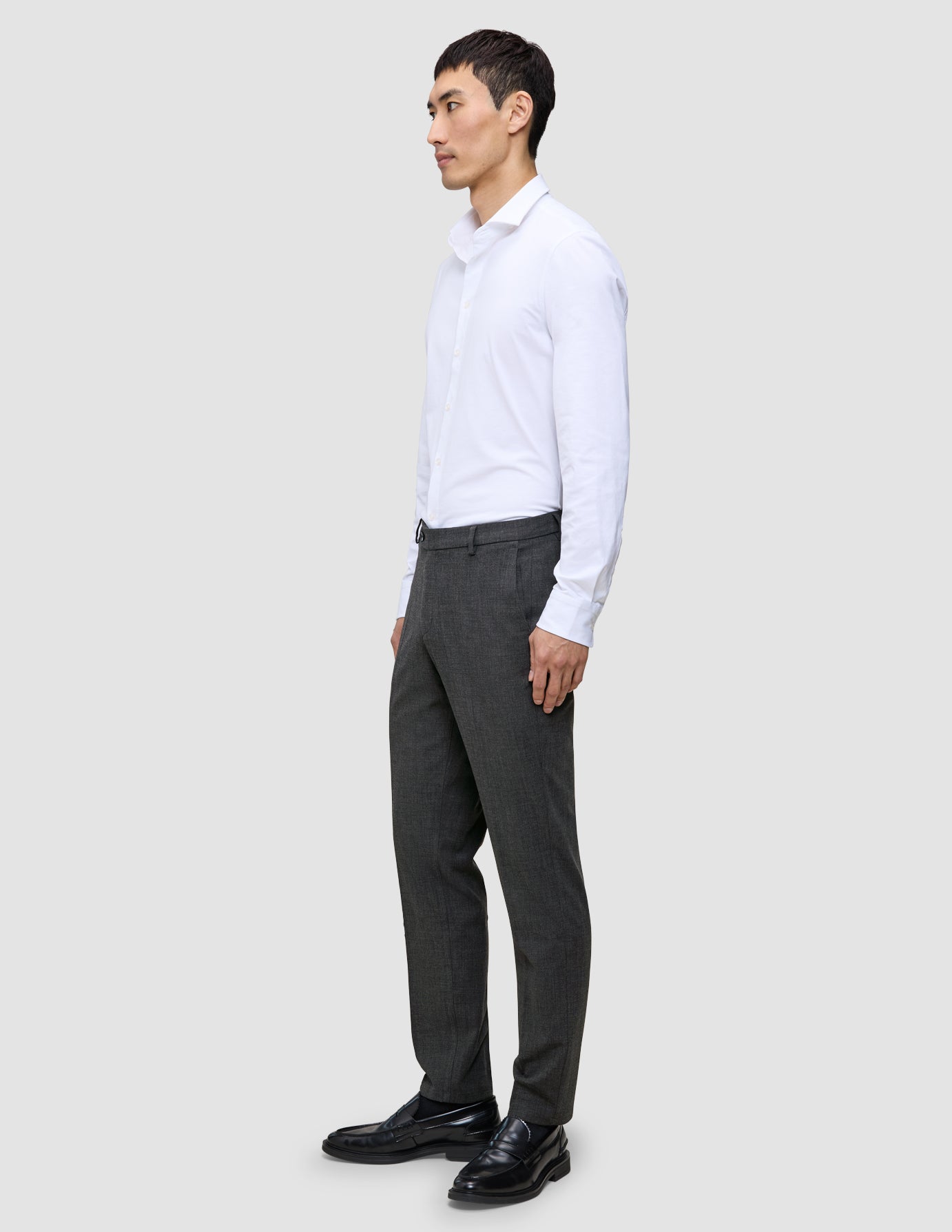 En mann i en hvit skjorte med knapper og grå Motion Suit-bukser i Regular Pepper Grey står sett i profil.