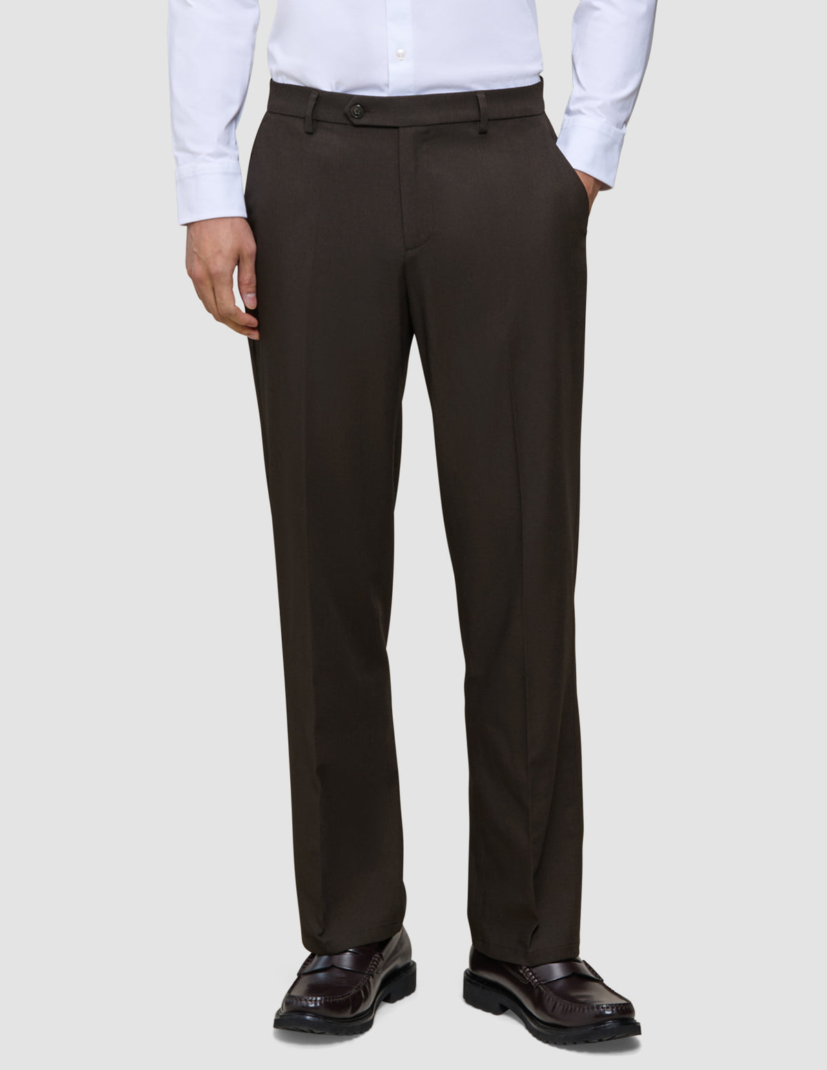 En person har på seg brune Motion Suit-bukser i modellen Relaxed Cognac, en hvit skjorte med knapper og mørke skinnloafers.