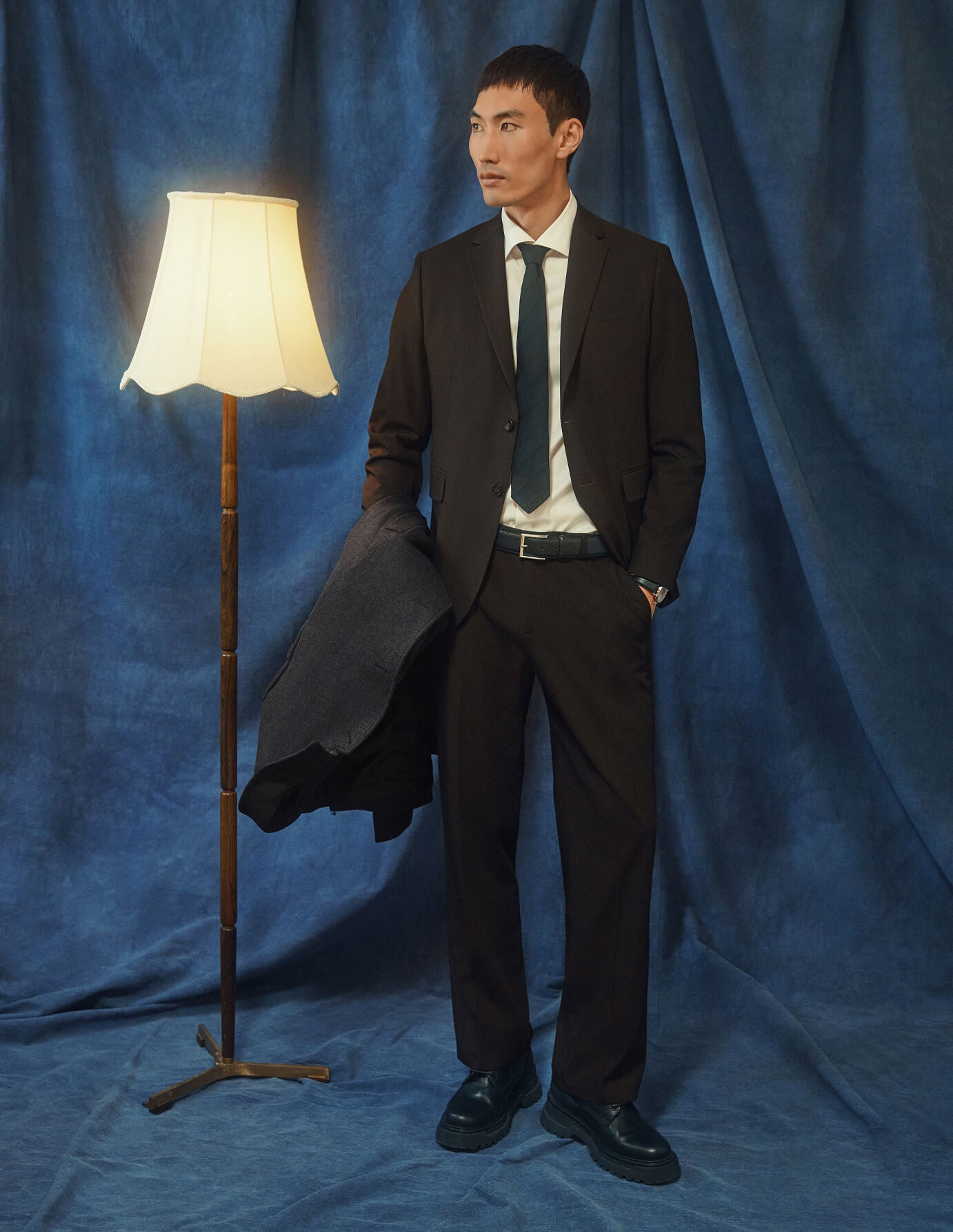 En mann kledd i Motion Suit Pants Relaxed Cognac, en hvit skjorte og et slips, som står ved siden av en gulvlampe.