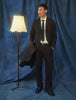 En mann kledd i Motion Suit Pants Relaxed Cognac, en hvit skjorte og et slips, som står ved siden av en gulvlampe.