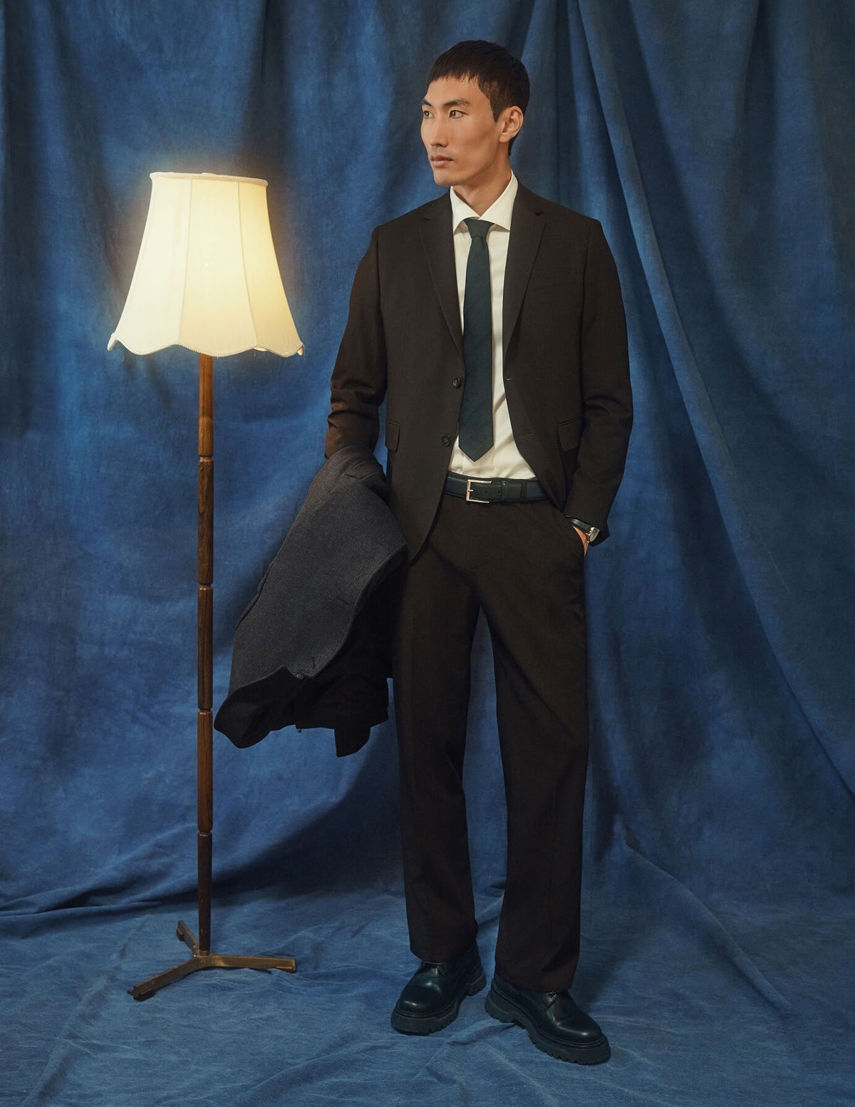 En mann kledd i Motion Suit Pants Relaxed Cognac, en hvit skjorte og et slips, som står ved siden av en gulvlampe.