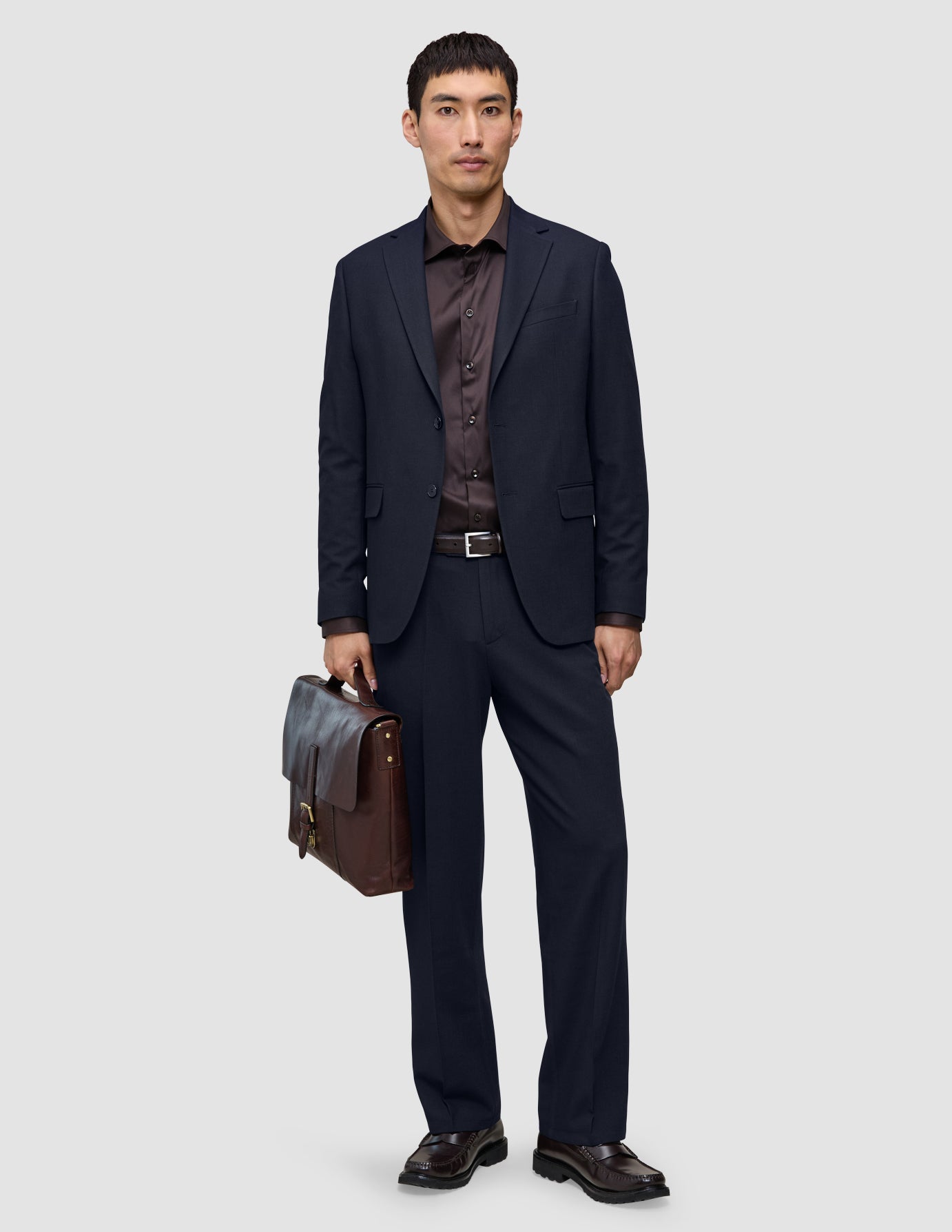 En mann i marineblå Motion Suit Pants Relaxed Midnight Blue, mørk skjorte og brune loafers, som holder en brun skinnkoffert.