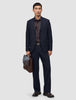 En mann i marineblå Motion Suit Pants Relaxed Midnight Blue, mørk skjorte og brune loafers, som holder en brun skinnkoffert.