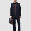 En mann i marineblå Motion Suit Pants Relaxed Midnight Blue, mørk skjorte og brune loafers, som holder en brun skinnkoffert.