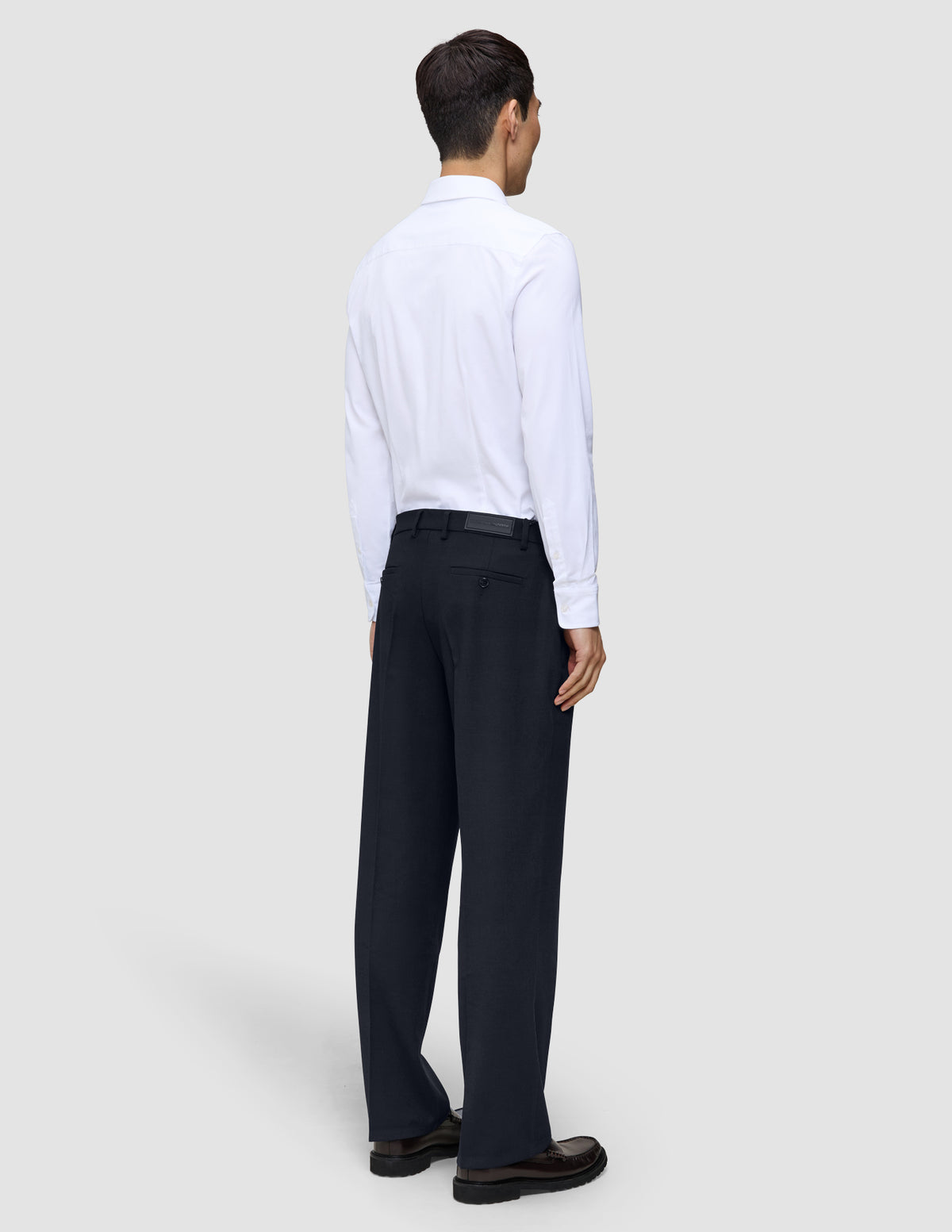 En person sett bakfra, kledd i en hvit skjorte med knapper og marineblå Motion Suit-bukser i avslappet passform i fargen Midnight Blue.