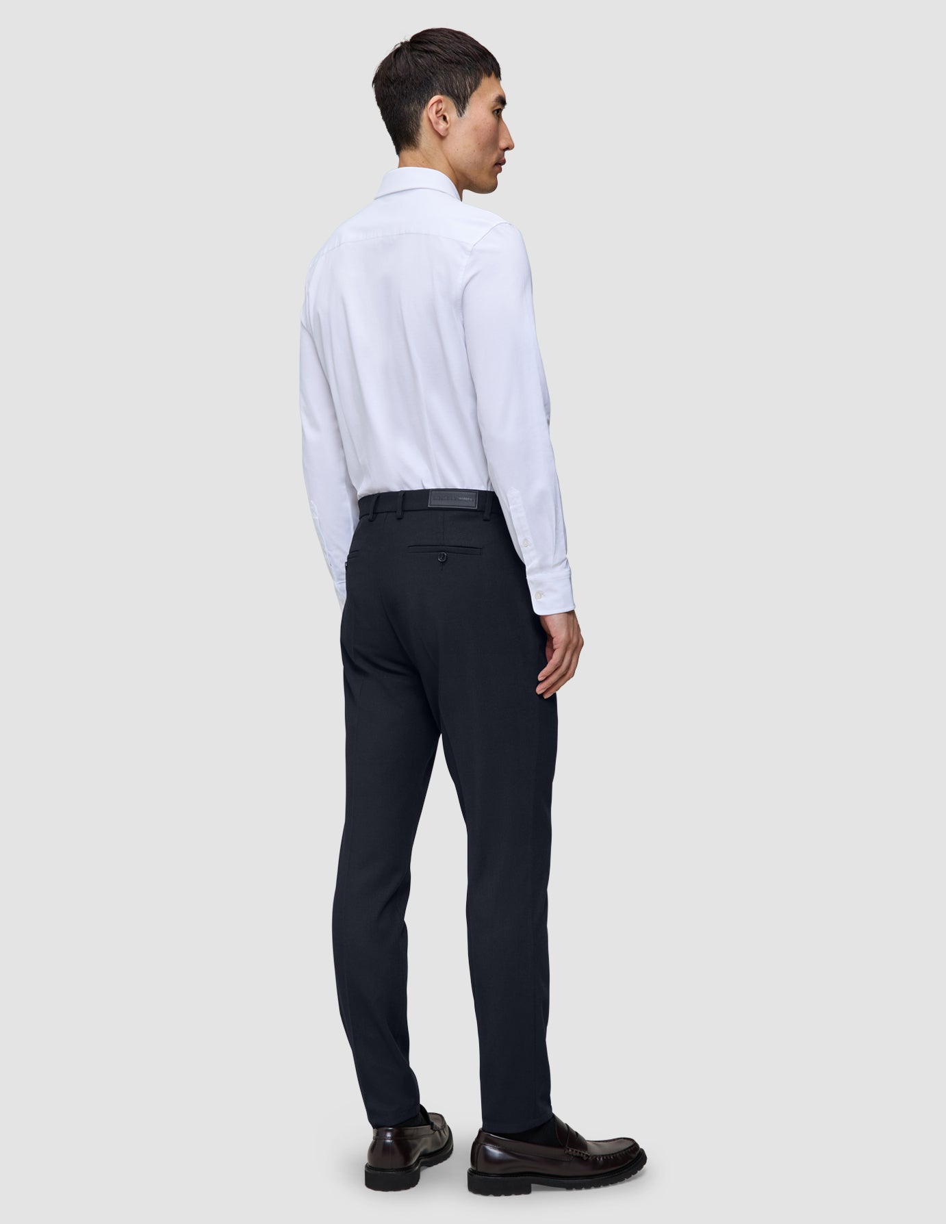 En mann sett bakfra, kledd i en hvit skjorte, Motion Suit Pants Slim i midnattsblått og mørke skinnloafers.