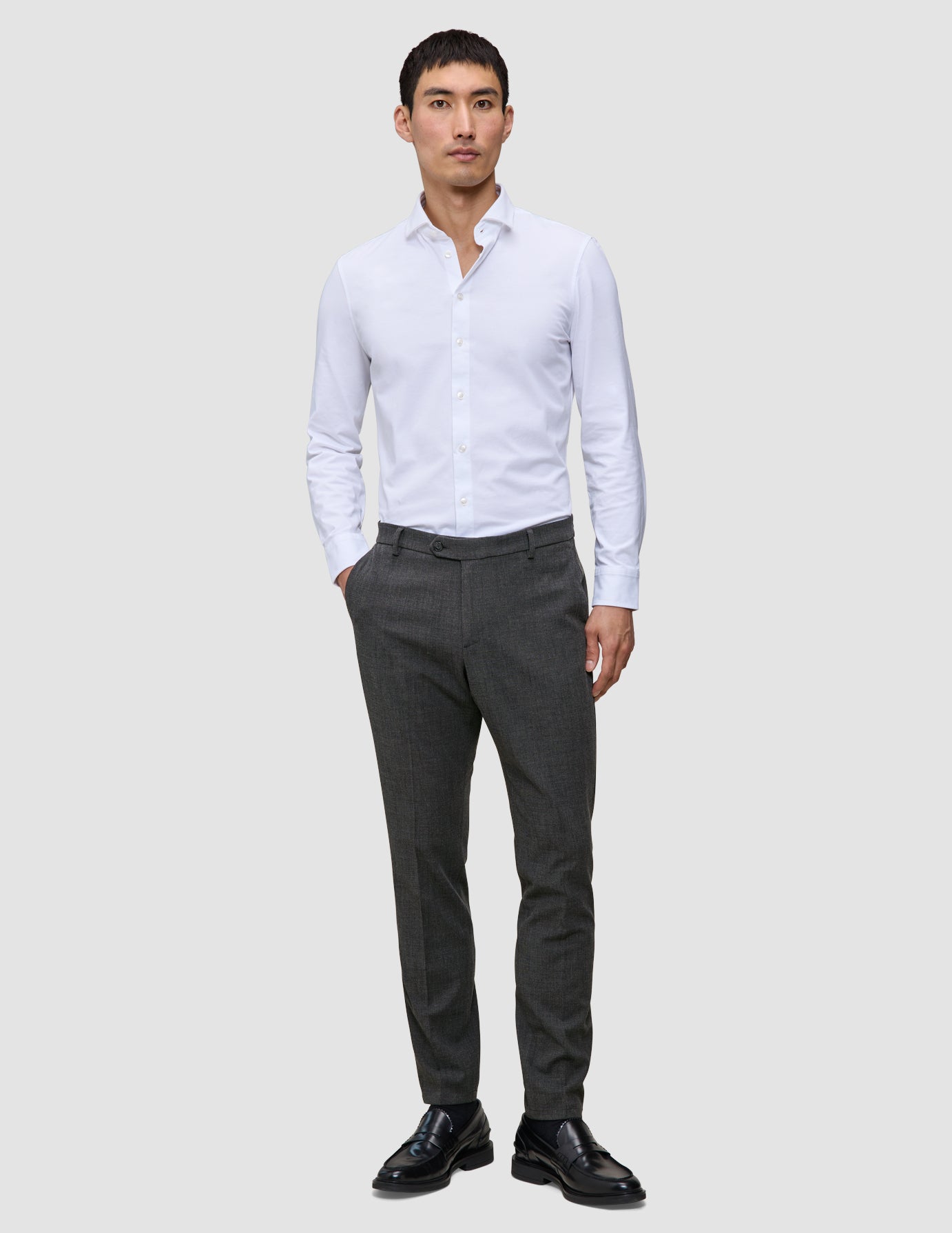 En mann kledd i en hvit skjorte med knapper, Motion Suit Pants Slim Pepper Grey og svarte loafers.
