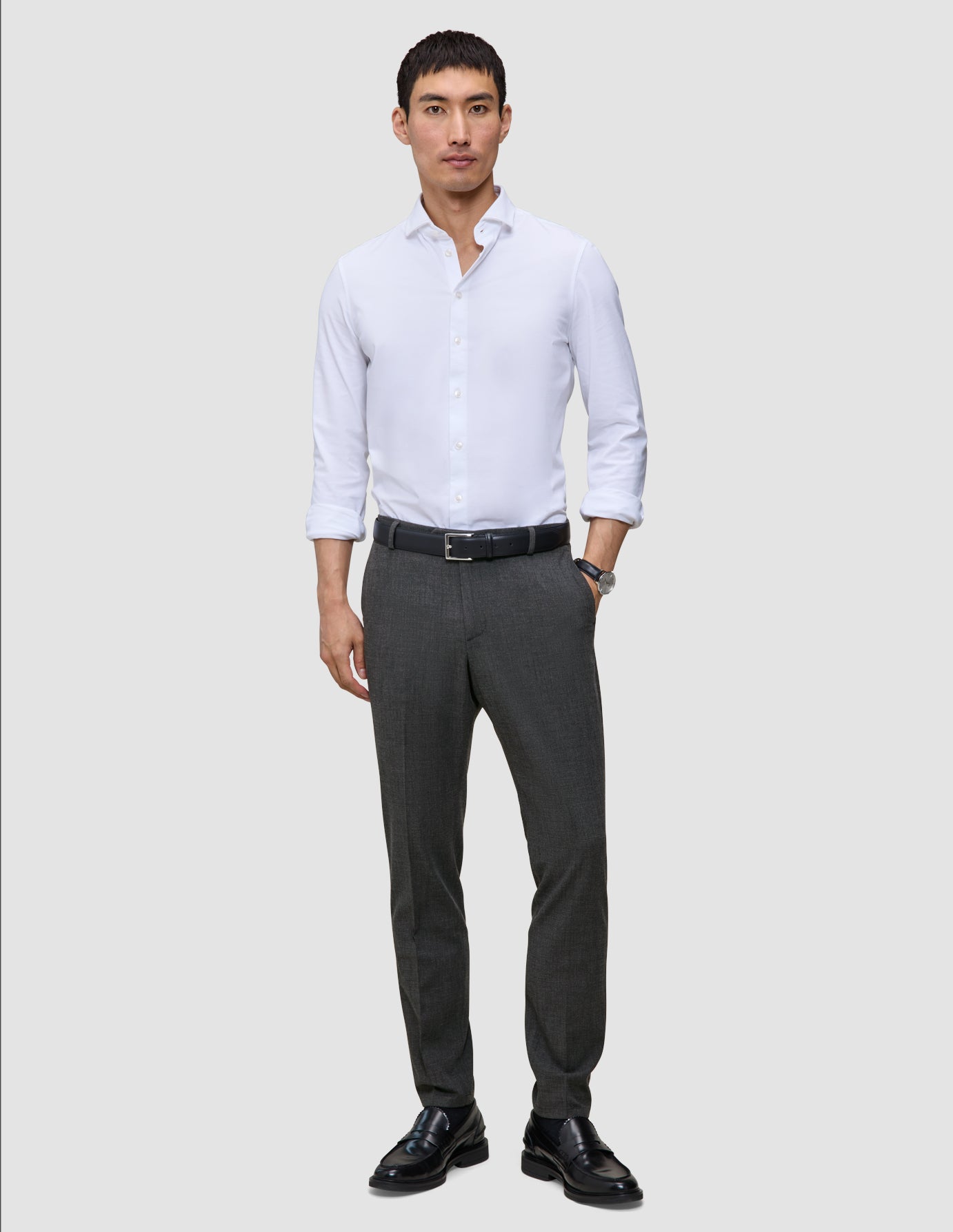 En mann i en hvit skjorte med knapper og Motion Suit-bukser i fargen Slim Pepper Grey, kombinert med et svart belte og loafers.