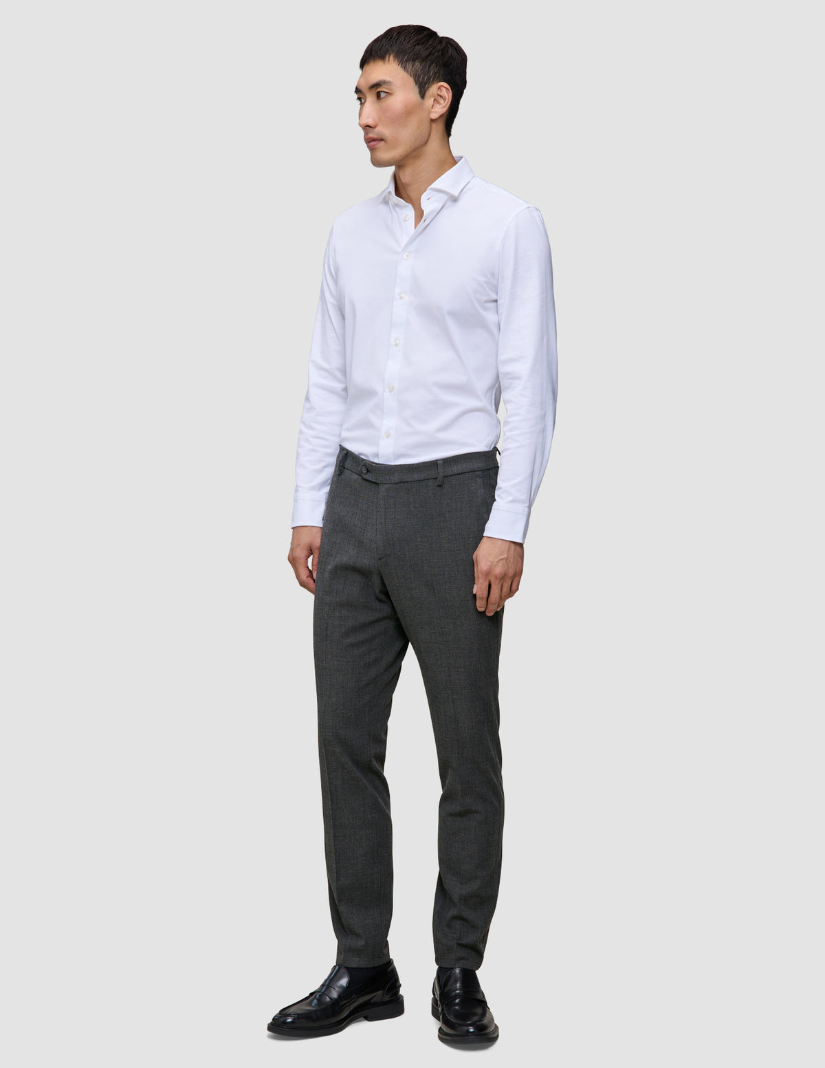 En mann i en hvit skjorte med knapper, Motion Suit-bukser i Slim-modell i fargen Pepper Grey og svarte dressko.