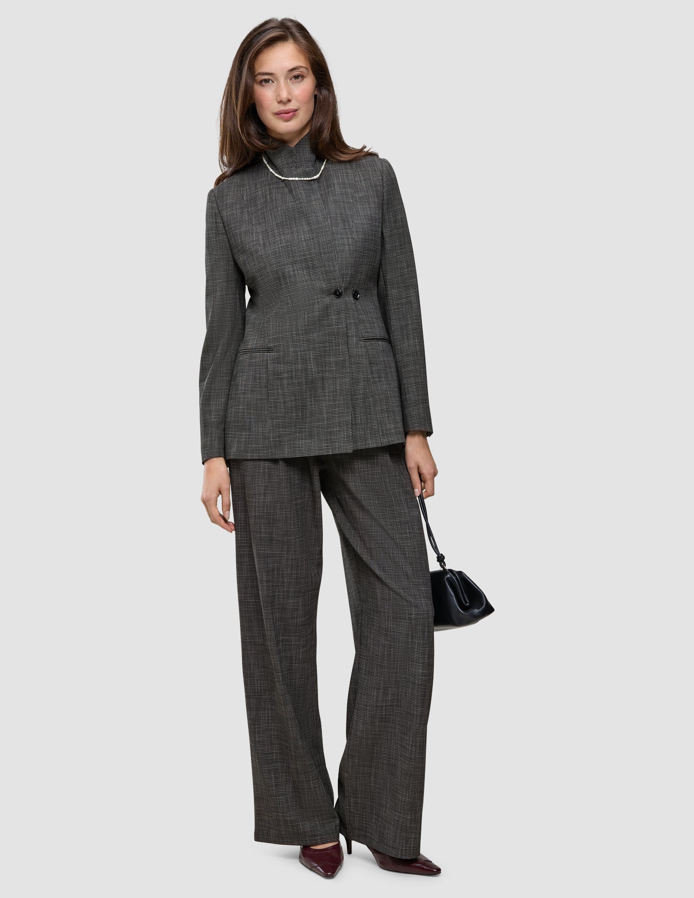 En person i dress med en uttalelse, kledd i en grå rutete blazer og matchende bukser med vide ben, som holder en svart håndveske.