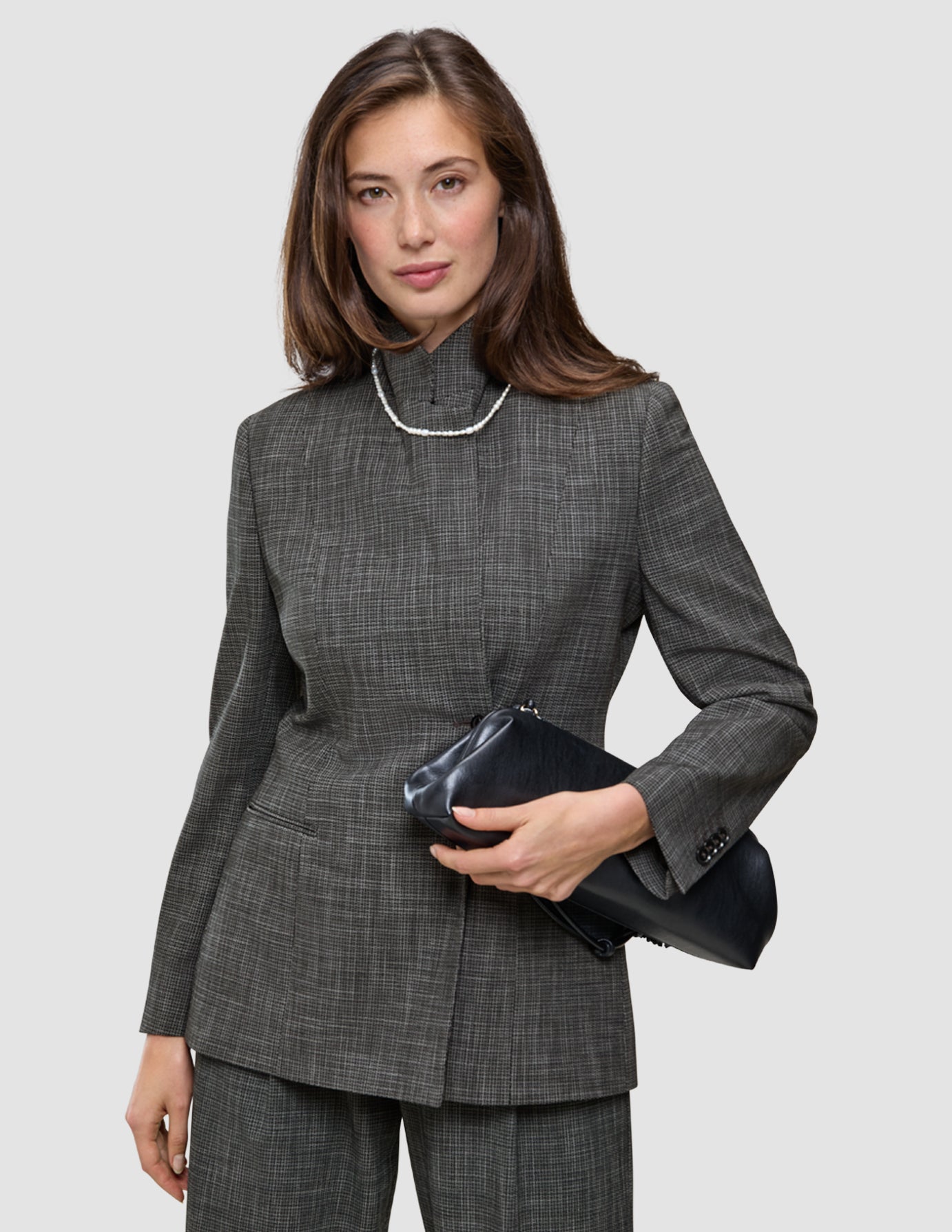 En kvinne iført en drakt med et iøynefallende mønster i grå rutemønster, som holder en svart clutch og har på seg et perlekjede.