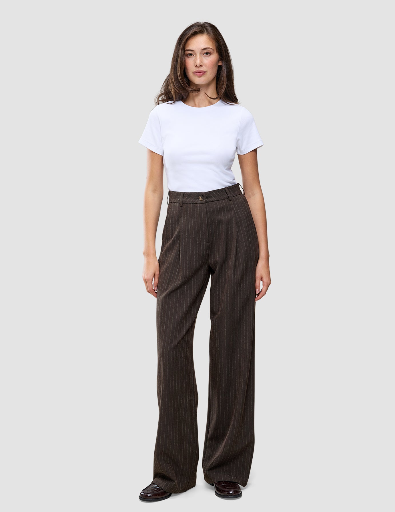 En kvinne har på seg en hvit T-skjorte og Serene Pants Wide Walnut Herringbone, som har et minimalistisk mørkebrunt design med smale striper.