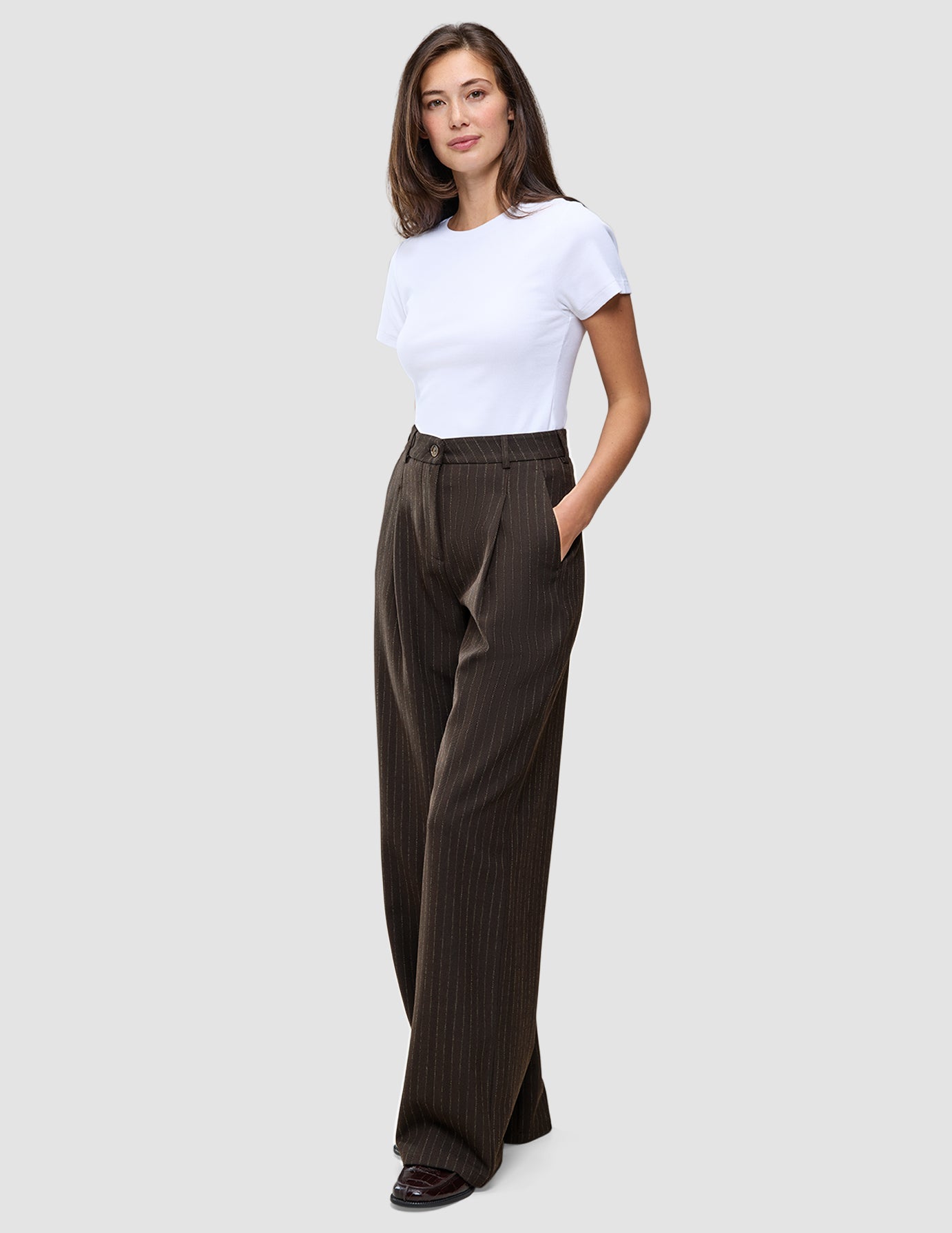 En person har på seg en hvit T-skjorte og Serene Pants Wide Walnut Herringbone – en buksemodell med høy midje, vide ben og nålestriper.