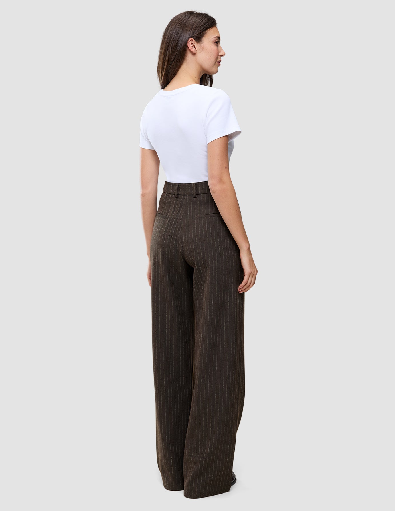 En kvinne sett bakfra, kledd i en hvit T-skjorte og Serene Pants Wide Walnut Herringbone med en diskret vertikal nålestripe.