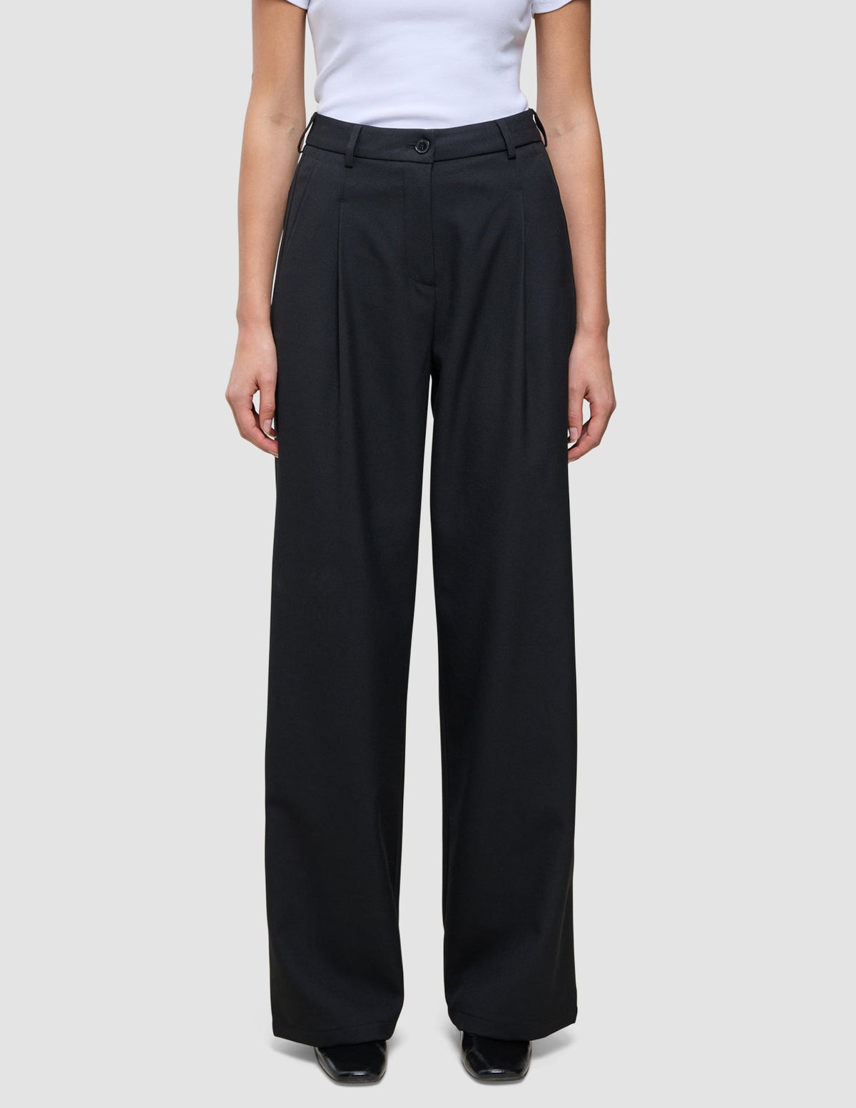 En person som har på seg de svarte Serene Pants Wide Black-buksene med plissering foran og en hvit T-skjorte i skandinavisk stil.