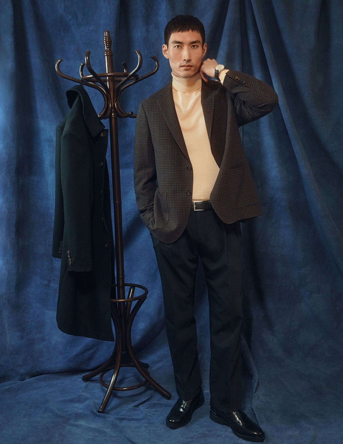 En mann i en mønstret blazer og en kremfarget turtleneck har på seg Tech Wool Suit Pants Relaxed Fit Black ved siden av en klesstativ.