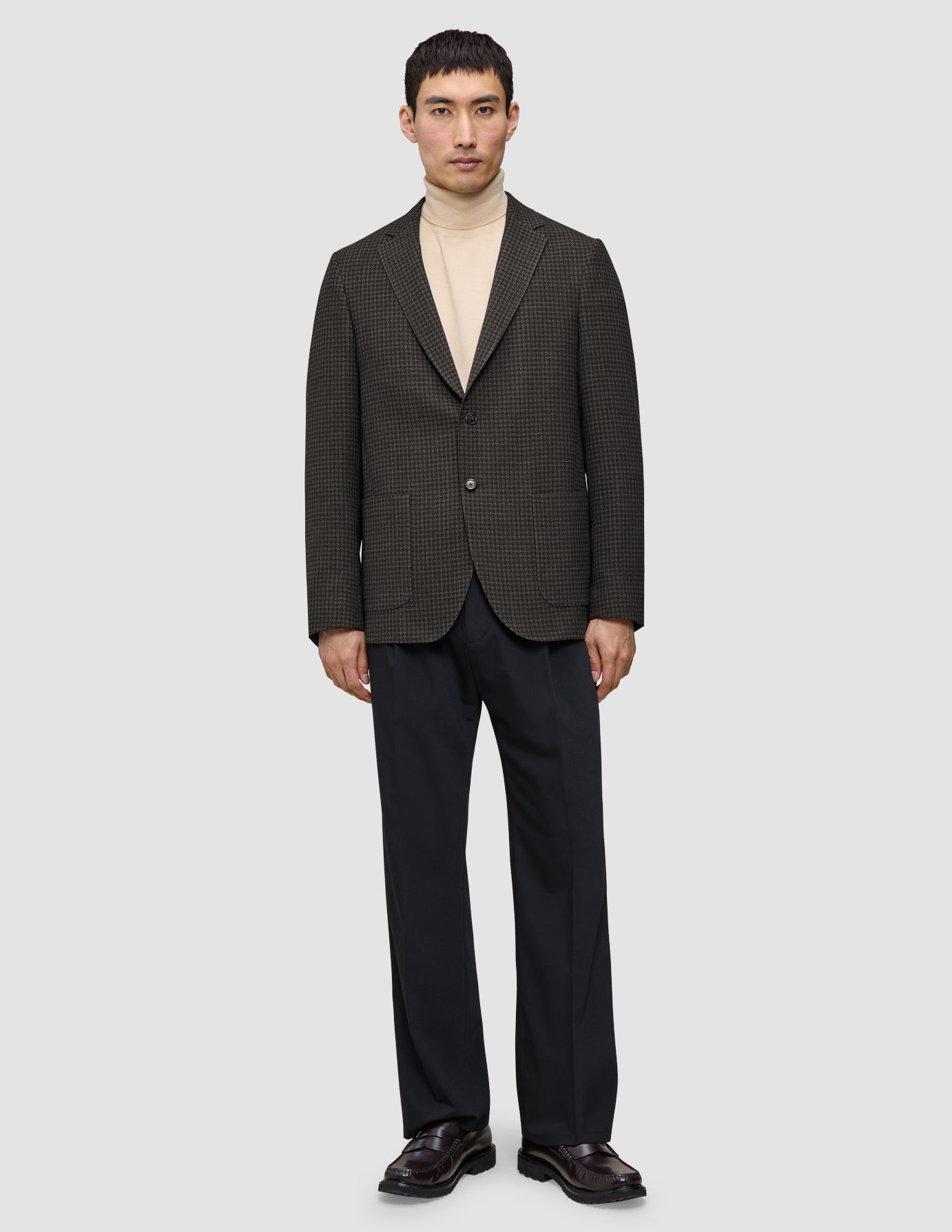 En mann har på seg en Timeless Wool Blazer Slim i mørkebrun rutemønster over en beige turtleneck og mørke bukser.