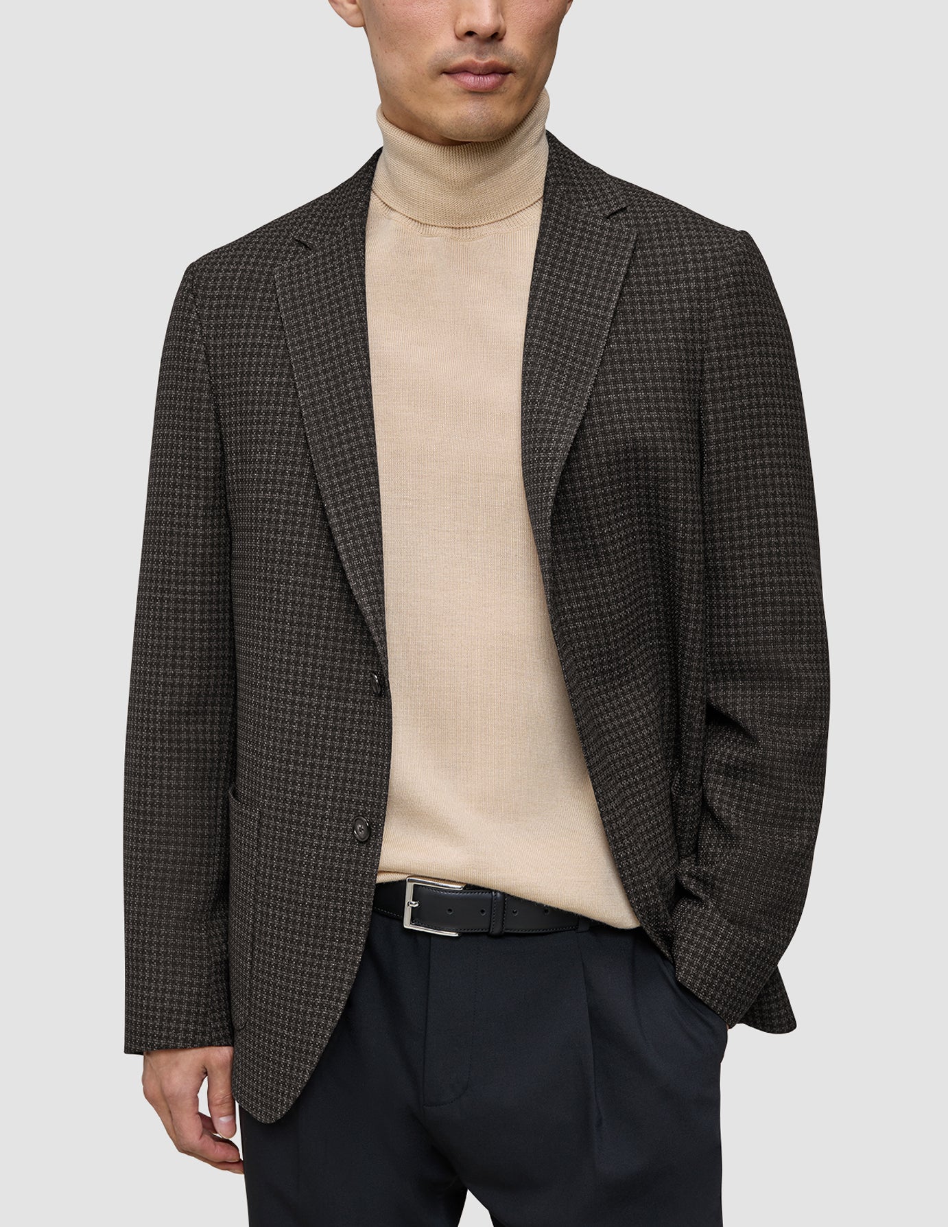 En person har på seg en Timeless Wool Blazer Slim i mørkebrun rutemønster over en beige turtleneck og svarte bukser.