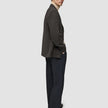 En mann kledd i en Timeless Wool Blazer Slim i mørkebrun rutemønster over en beige turtleneck og mørke bukser, som ser seg tilbake.