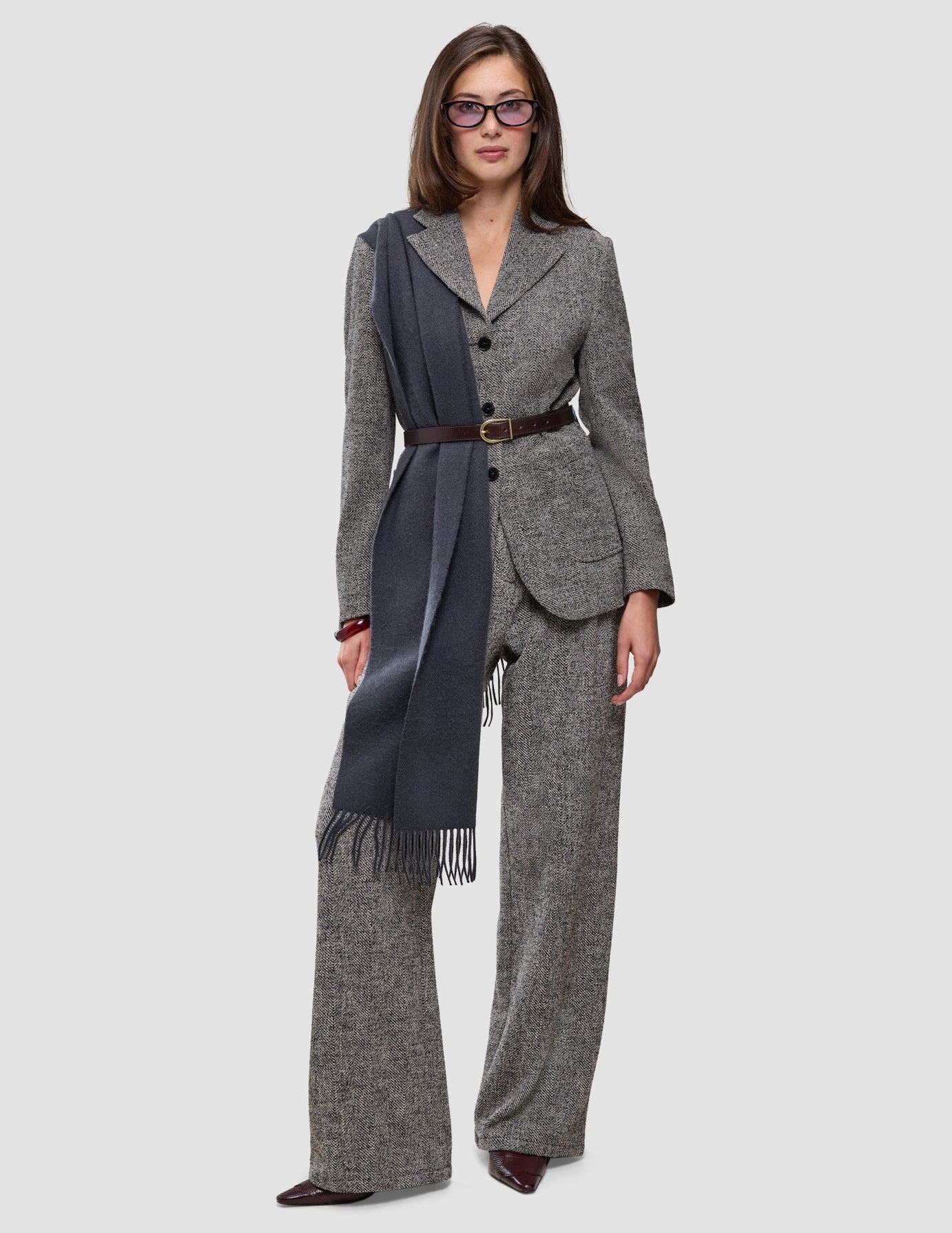 Kvinne i en grå blazer i bouclé med fiskebeinmønster, Ashryn-bukser for kvinner i bred modell med Domino-fiskebeinmønster, belte, briller og et langt grått skjerf.
